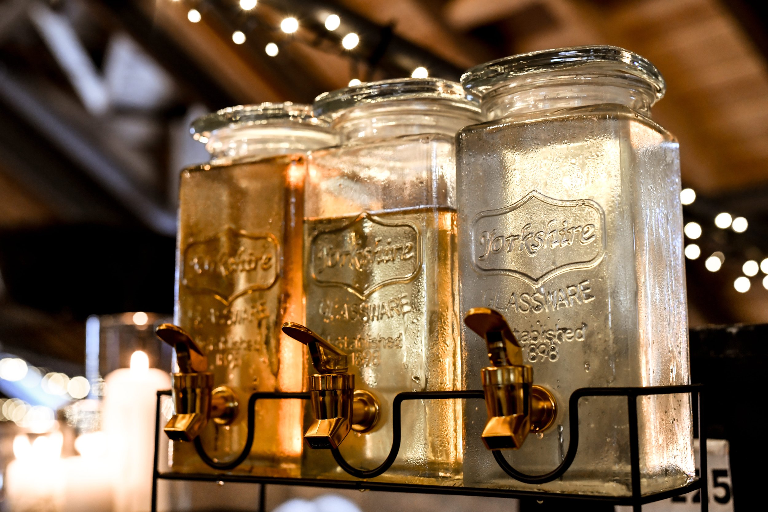 Des bocaux en verre contenant des boissons, types limonades maison, sont placés sur un support noir avec une ambiance chaleureuse et des lumières floues en arrière-plan.