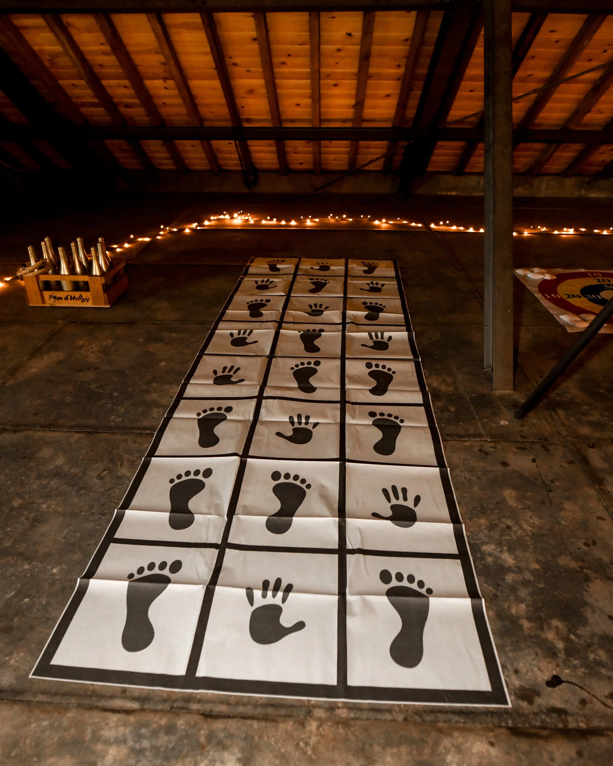 Une salle avec un sol en bois, décorée avec des guirlandes lumineuses. Au sol, un tapis avec des empreintes de pas de différentes tailles, utilisés pour une activité de jeu ou de danse. À gauche, un carton contenant des bouteilles en verre, et à droi
