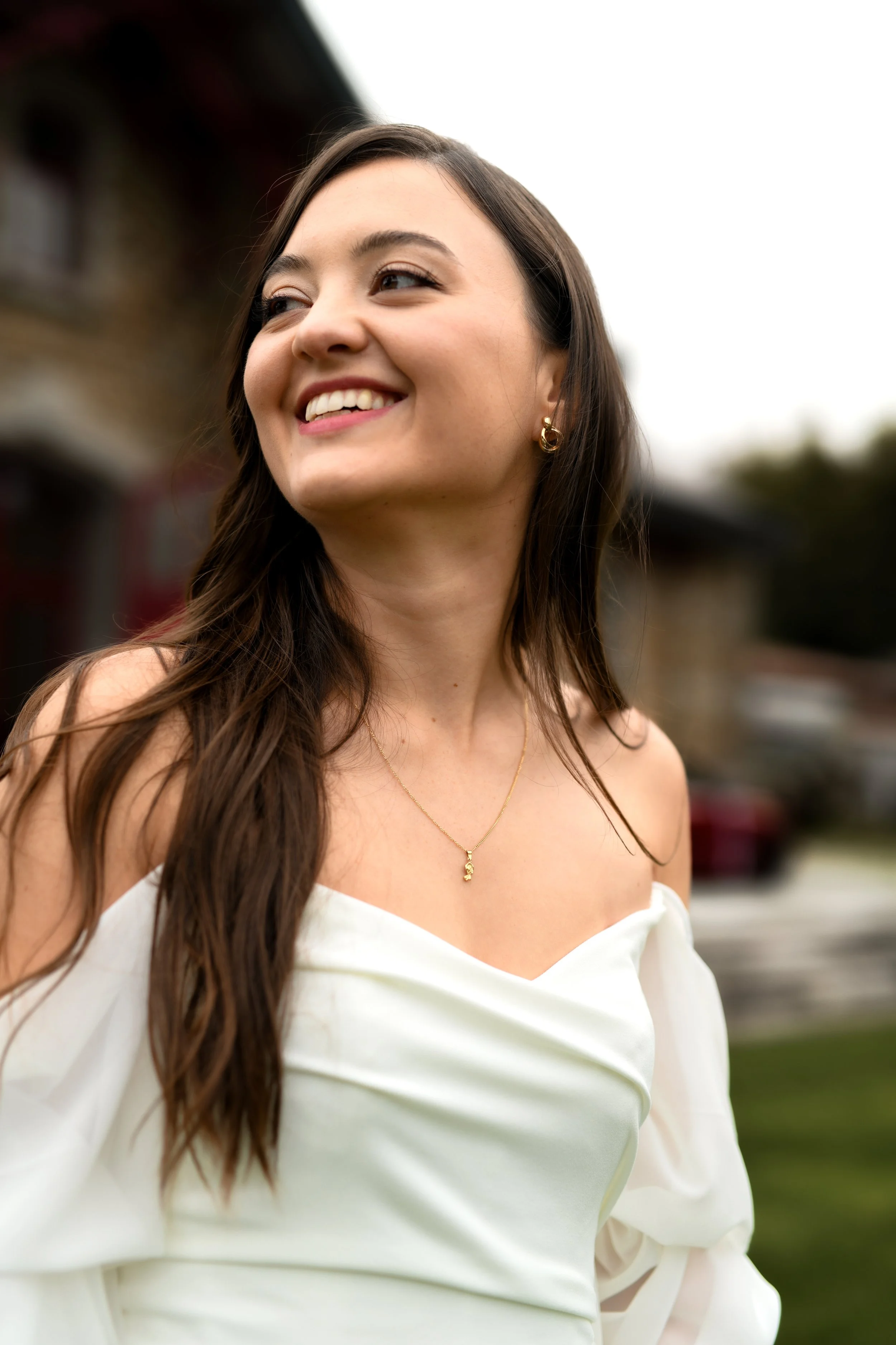 Jeune femme souriante portant une robe blanche et des bijoux en or, en extérieur.