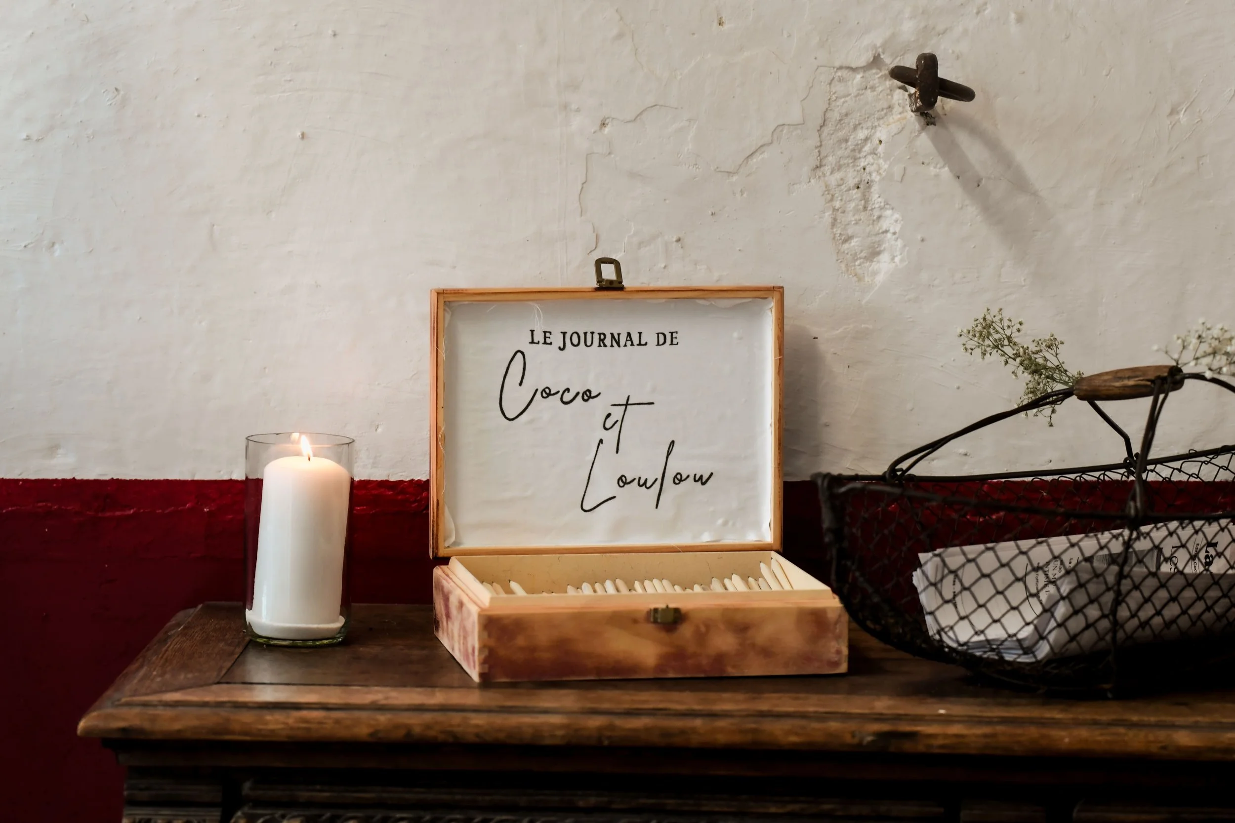 Une bougie allumée, une boîte en bois ouverte avec des blocs d'écriture, un panier en métal et une plante sèche sur une table en bois, contre un mur blanc avec une moitié rouge.