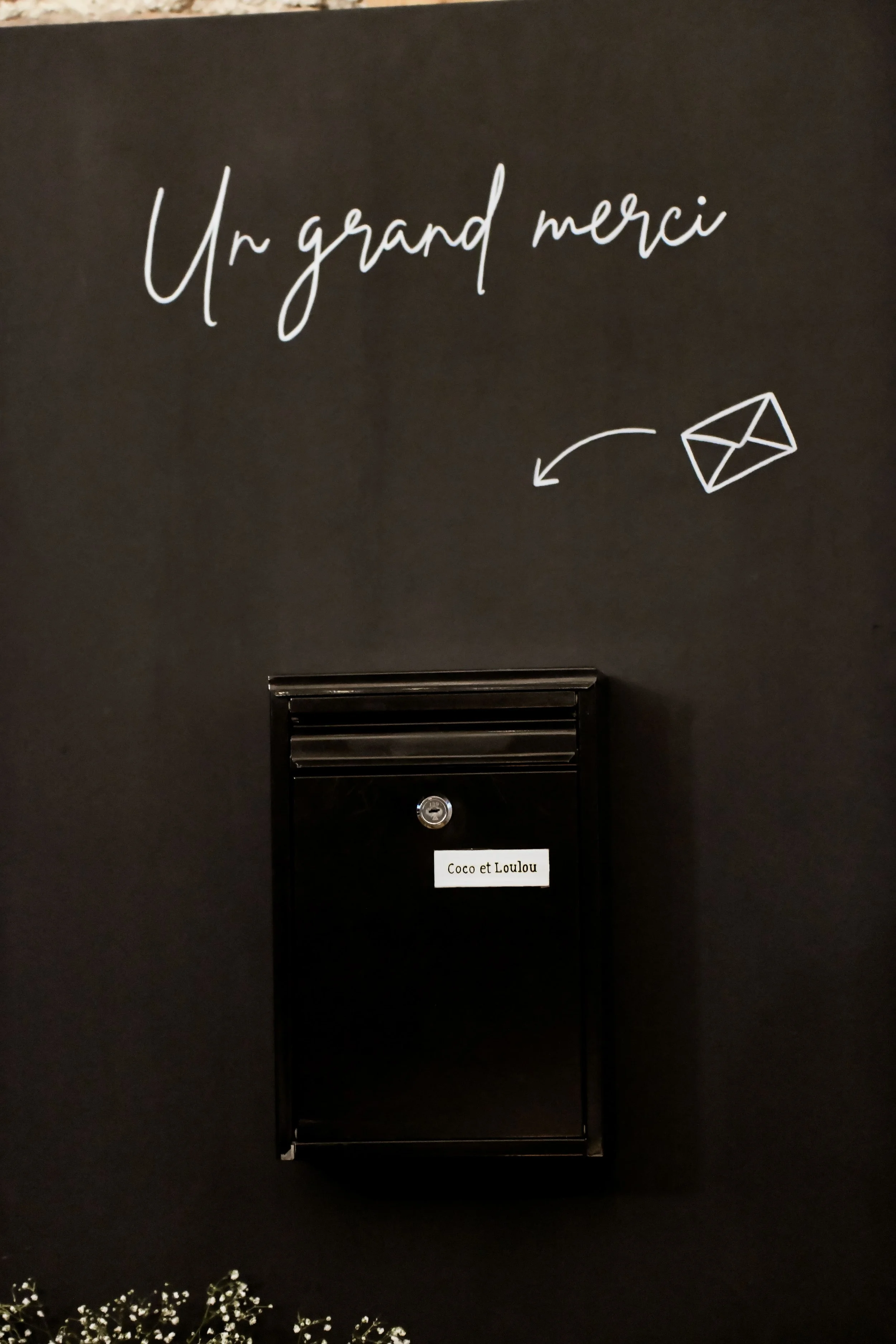 Une boîte aux lettres noire fixée sur un mur noir. Une étiquette blanche en dessous indique : Coco et Loulou. Au-dessus, il y a une écriture blanche qui dit : Un grand merci, avec un dessin d'une enveloppe à côté.