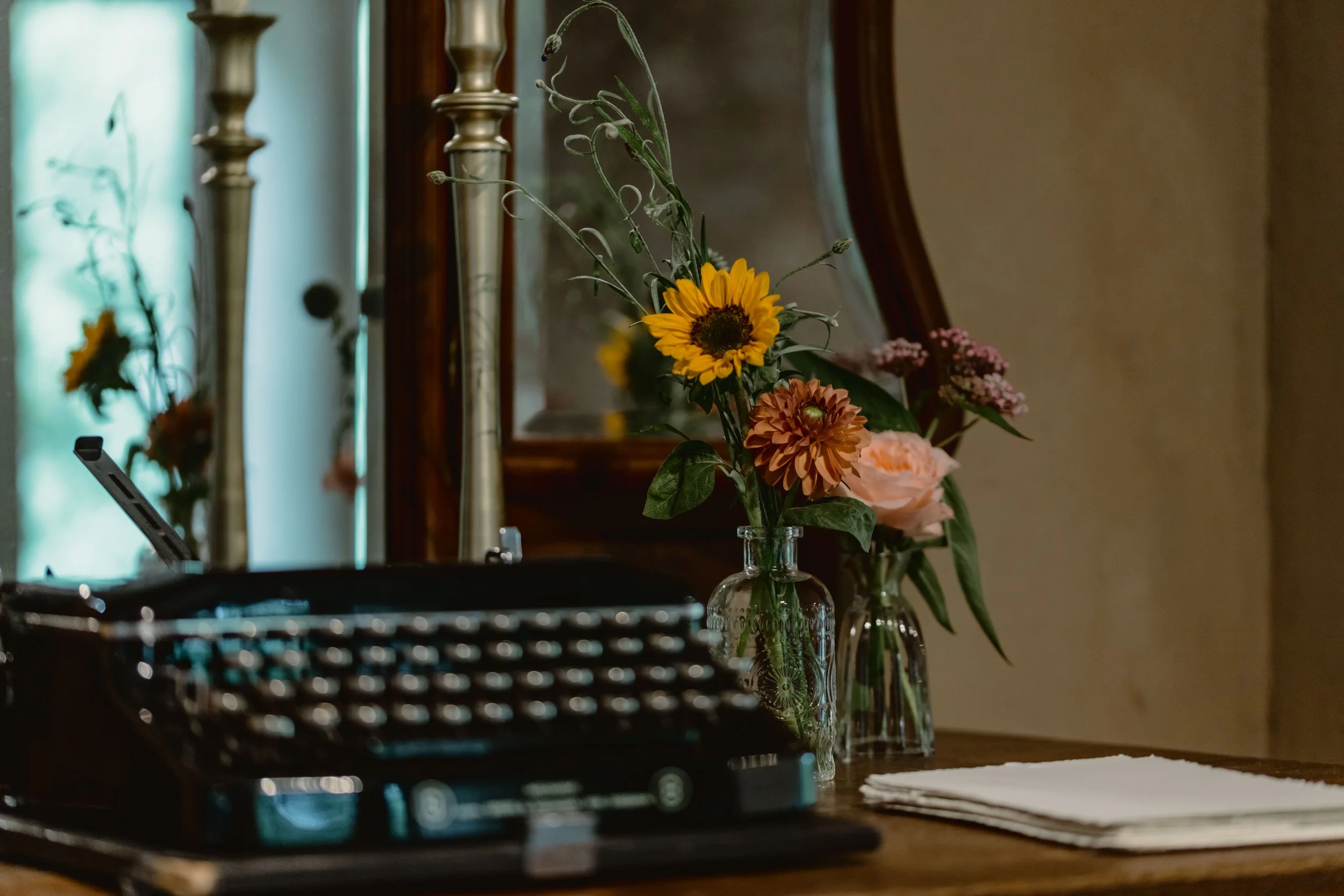 Un bouquet de fleurs, principalement des tournesols, dans un vase en verre placé sur une table en bois. En arrière-plan, un miroir et deux bougeoirs en métal avec des bougies. Le décor est chaleureux et traditionnel.