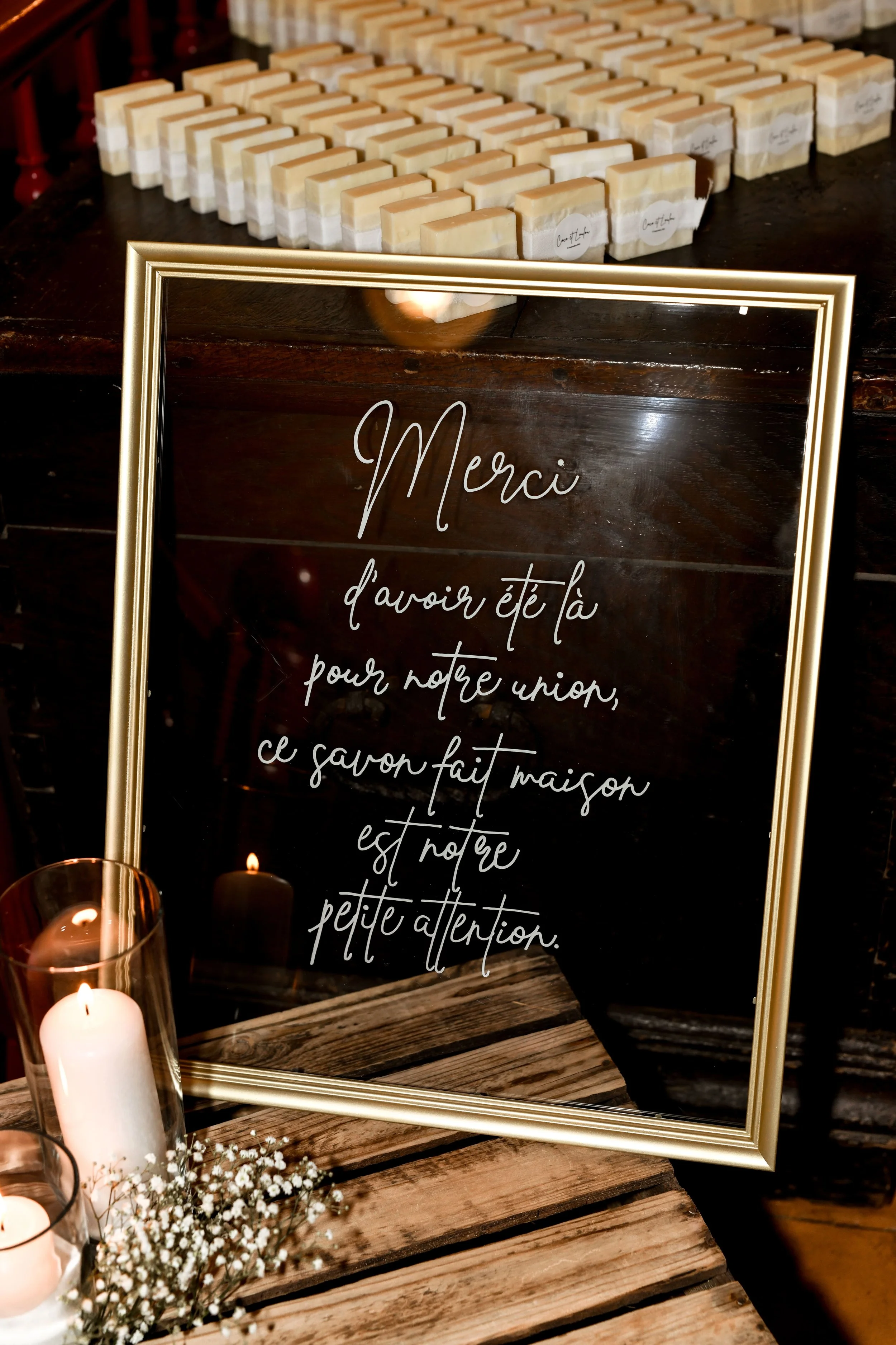 Un miroir rectangulaire avec un cadre doré, posée sur une table en bois, avec un message écrit en français : « Merci d'avoir été là pour notre union, ce savon fait maison est notre petite attention ». Derrière le miroir, il y a des savons en formes r