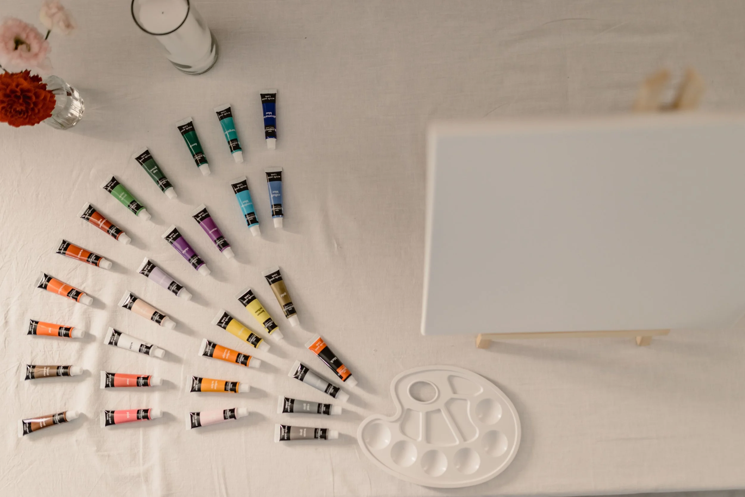 Une palette de peinture avec plusieurs tubes de peinture de différentes couleurs, une table avec des vases de fleurs et une toile blanche.