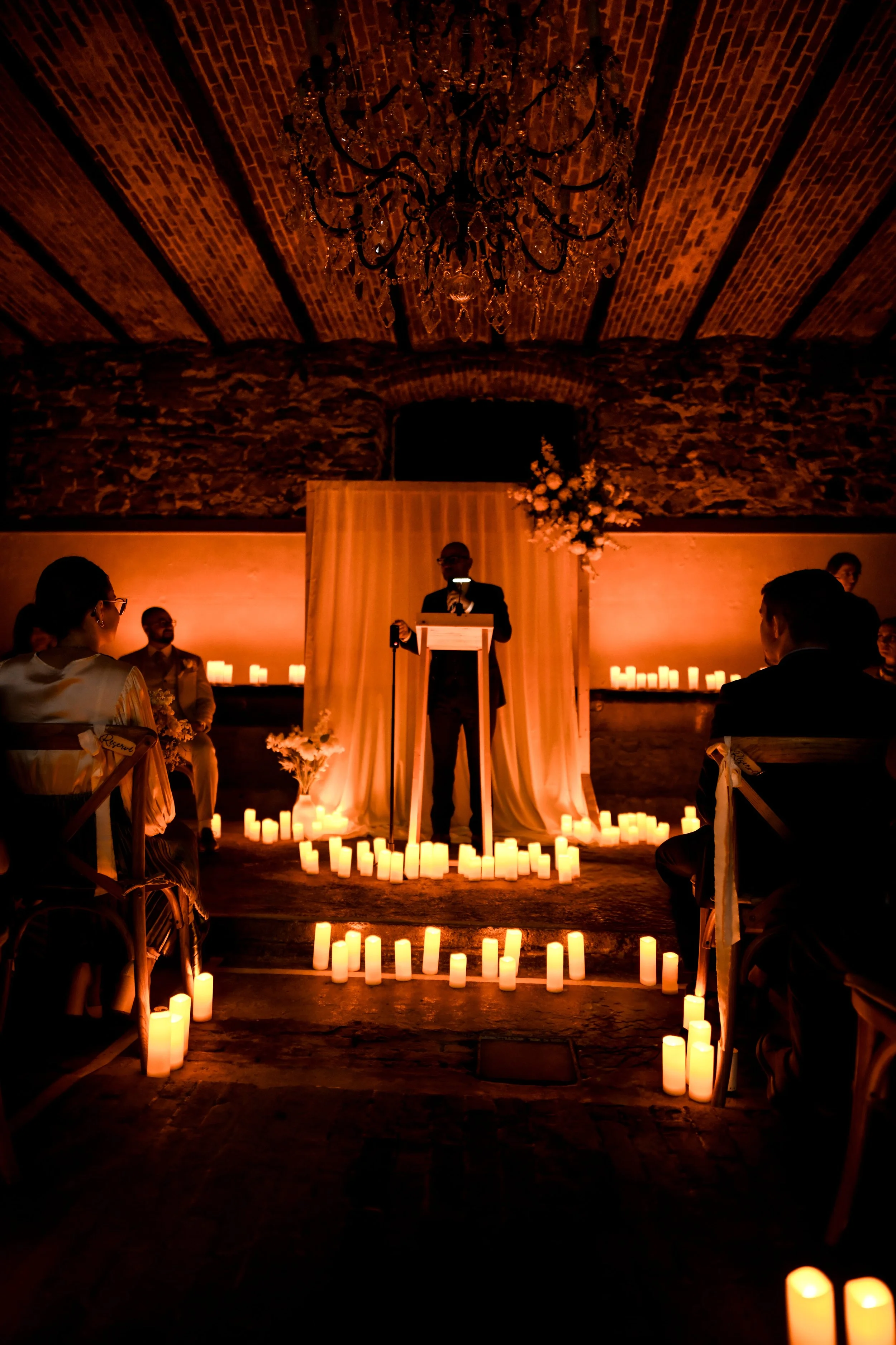 Une scène de mariage dans une salle avec une ambiance chaleureuse, éclairée par de nombreuses bougies. Un homme au centre parle derrière un podium sous un éclairage doux, avec un décor floral à côté. La pièce a un plafond en briques et une pierre anc