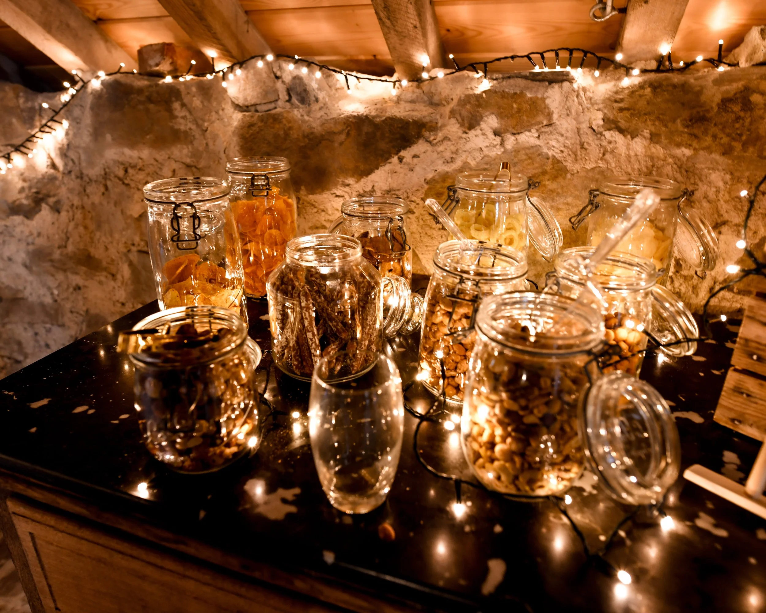 Verres en verre transparent remplis de bonbons variés derrière une surface en bois foncé, décorés avec des petites guirlandes lumineuses suspendues dans une cave ou un sous-sol en pierre et bois.