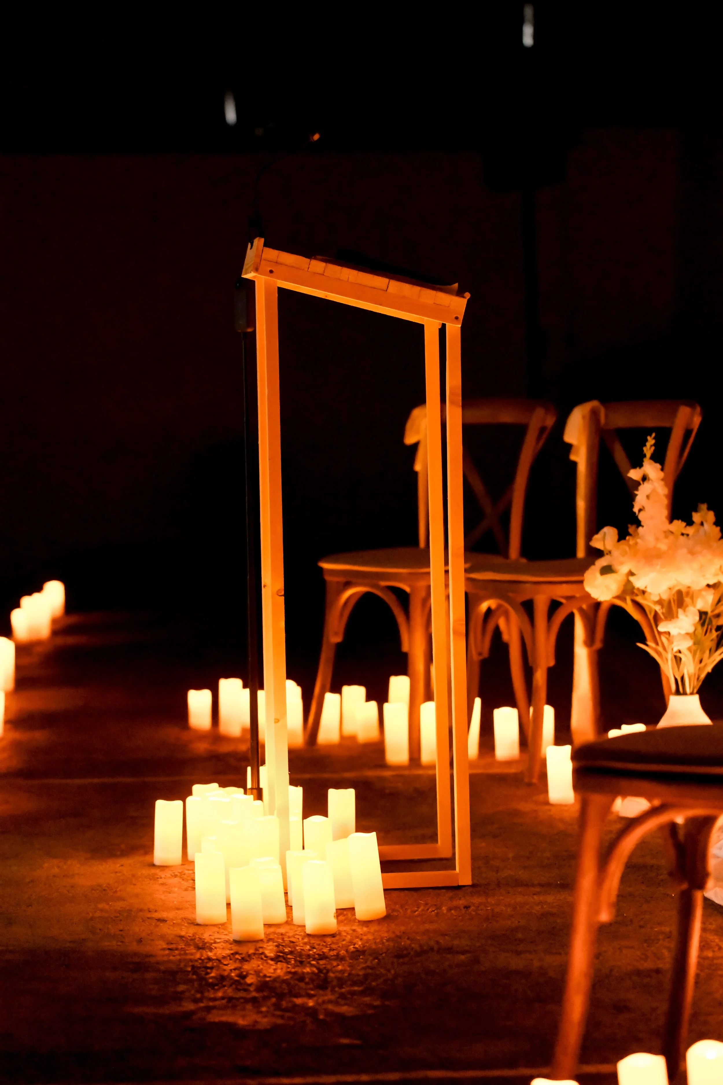 Décor intérieur avec bougies éclairant une scène, chaises en bois et fleurs, ambiance chaleureuse et romantique.