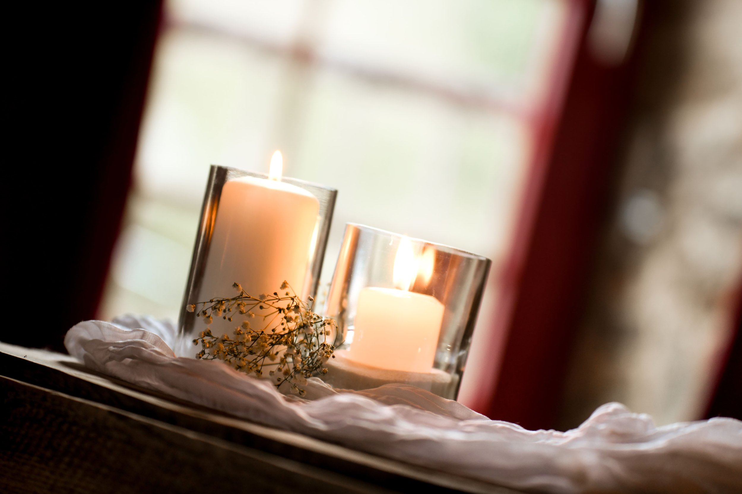 Une bougie dans un verre en verre avec des fleurs séchées à ses côtés, placée sur un linge blanc, vue à travers une ouverture floue.