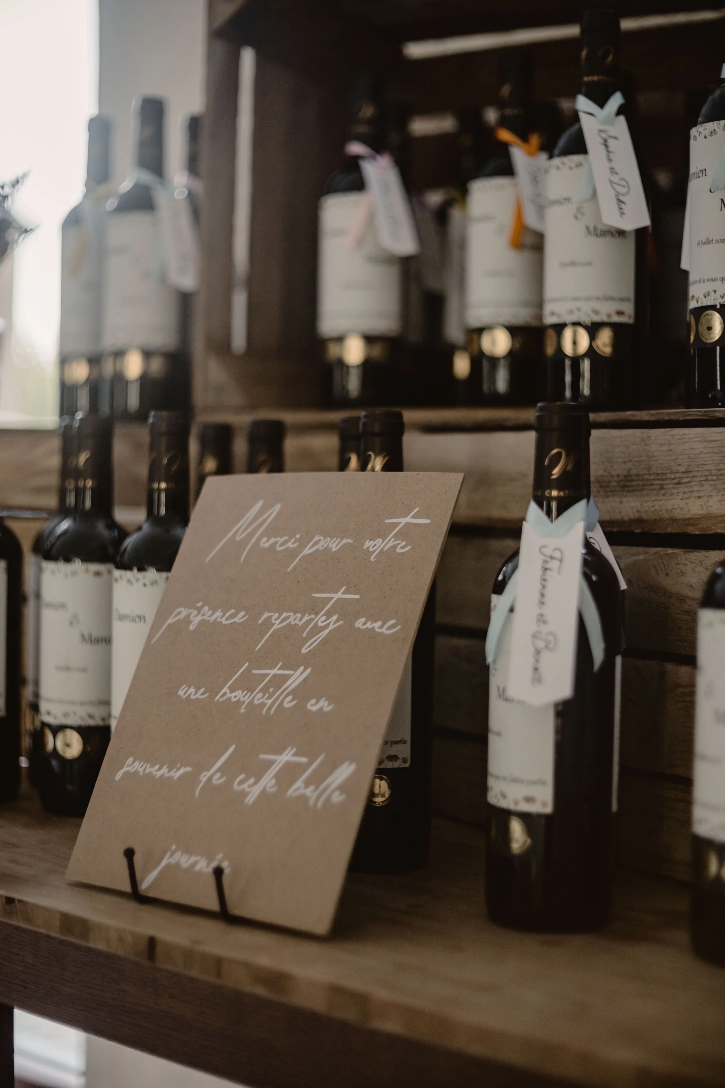 Les bouteilles de vin sont alignées sur une étagère en bois dans une cave ou un magasin. Un panneau en carton à l'avant porte une note manuscrite en français, exprimant la gratitude pour une visite ou un achat.