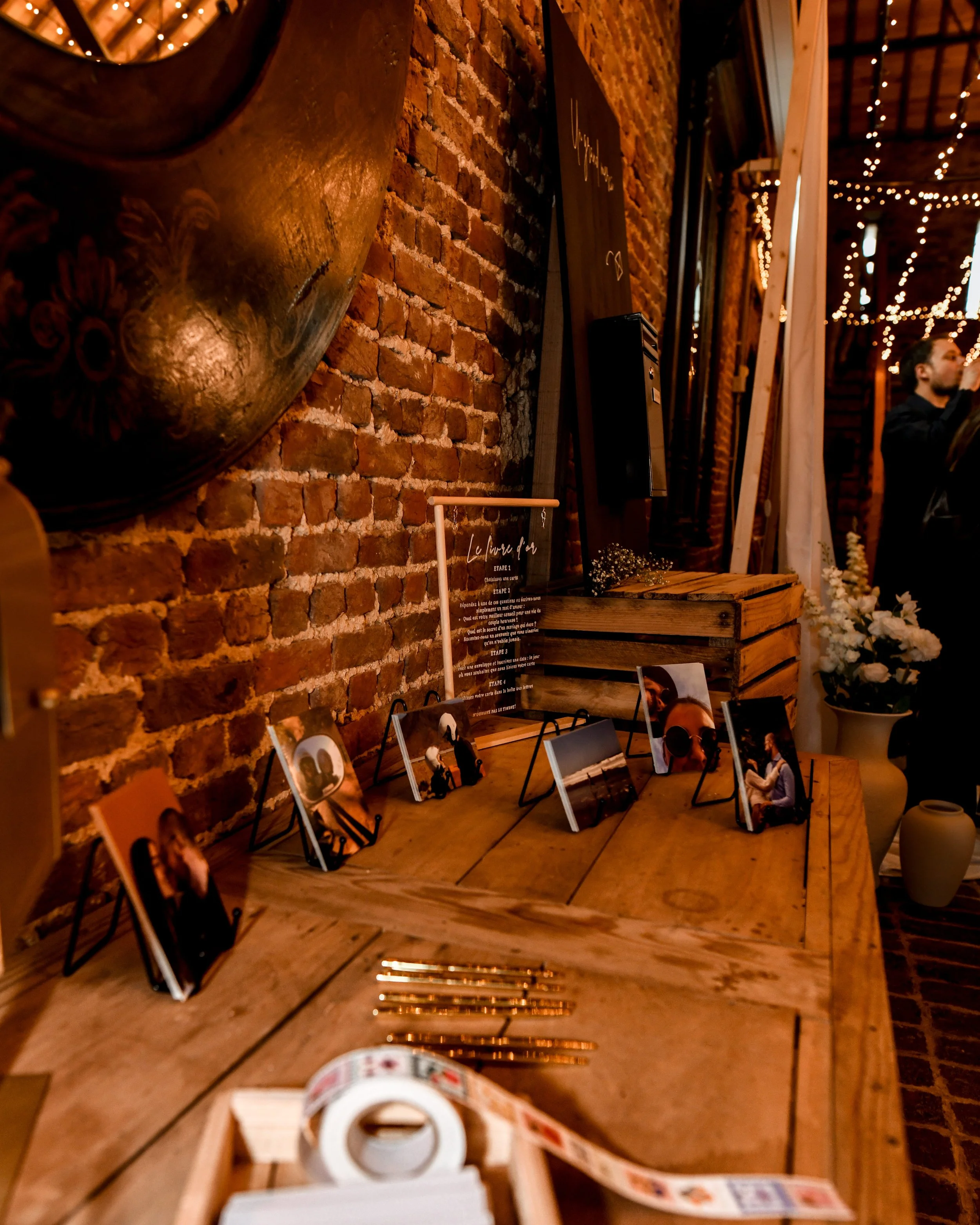 Décor intérieur avec mur en briques, guirlandes lumineuses suspendues, un tableau noir et des photographies sur une table en bois. Présence d'une personne à droite en arrière-plan.