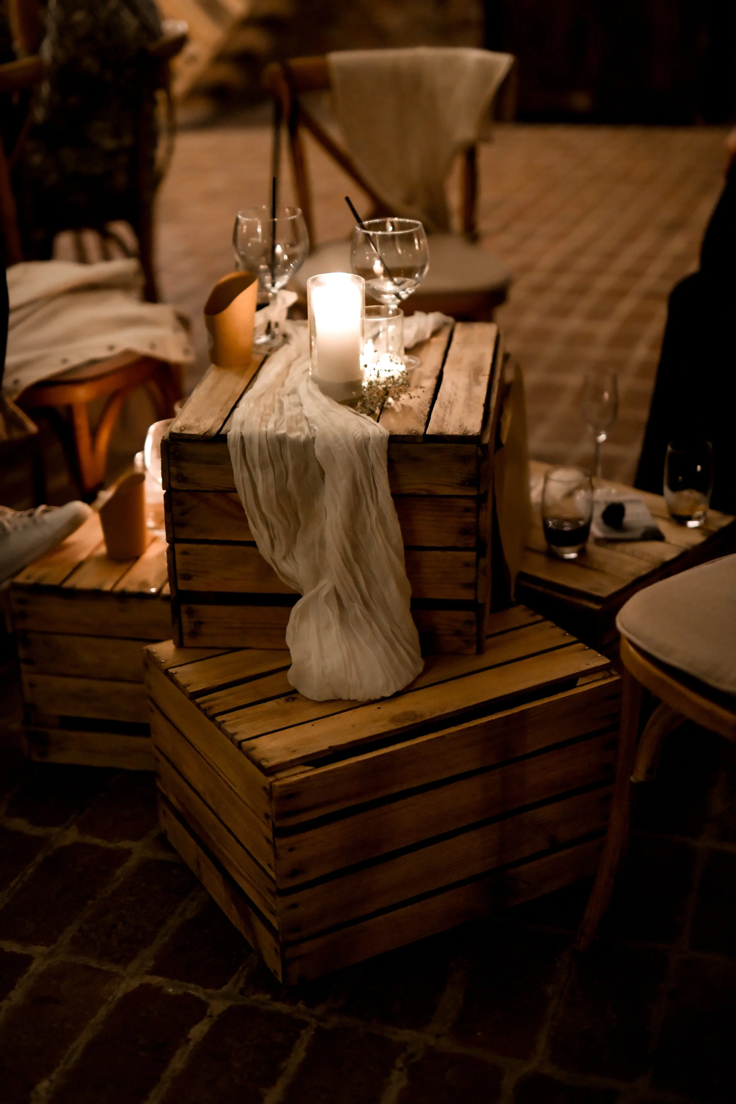 Une table en bois avec une bougie allumée, deux verres à vin, une serviette en tissu blanc, un pot en carton, et une petite décoration florale. Le décor semble être dans un lieu intérieur avec une ambiance chaleureuse et cosy.