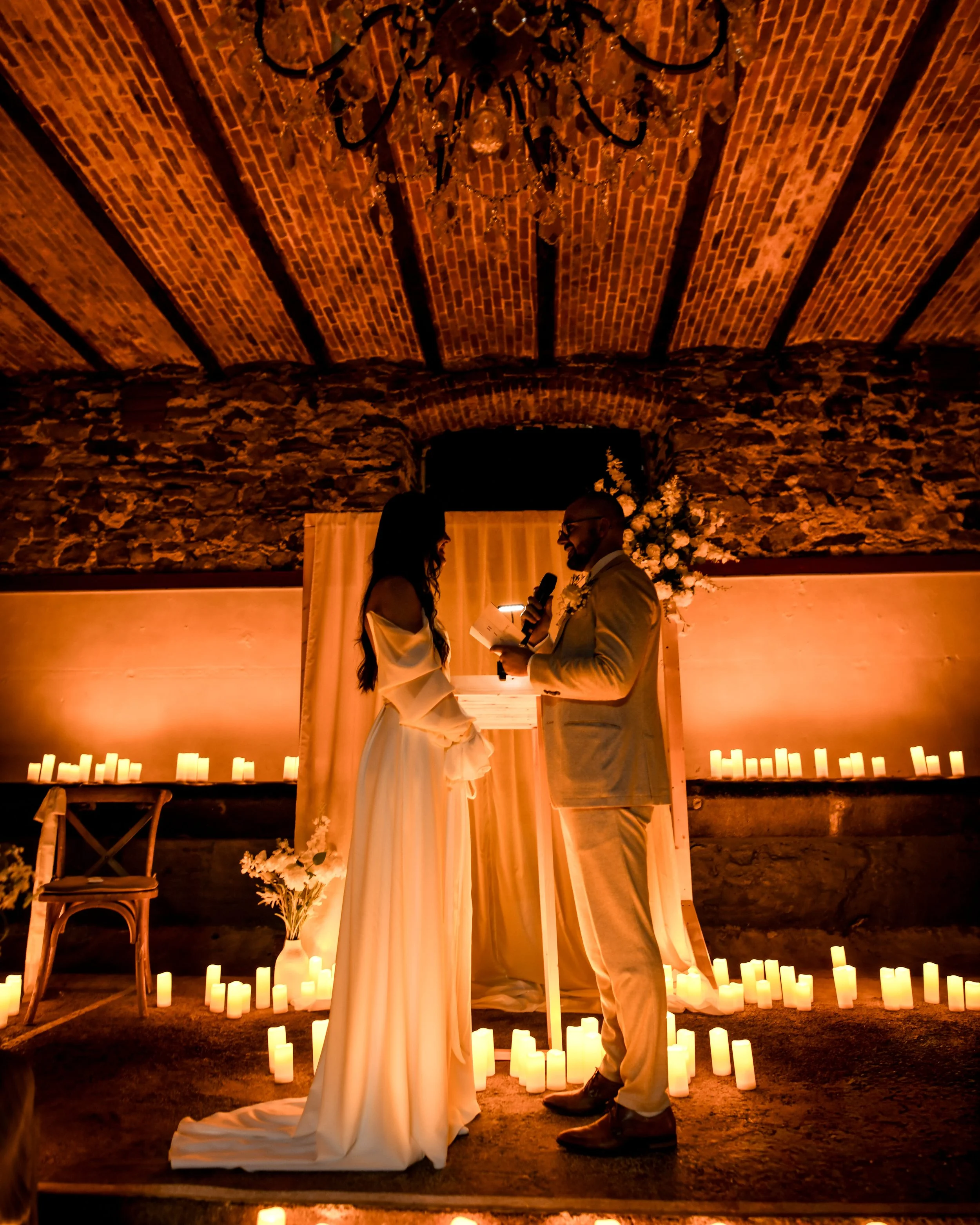 Un couple lors de la cérémonie de mariage, se tenant face à face, entouré de bougies sur le sol, dans un cadre rustique avec une arche et une décoration florale
