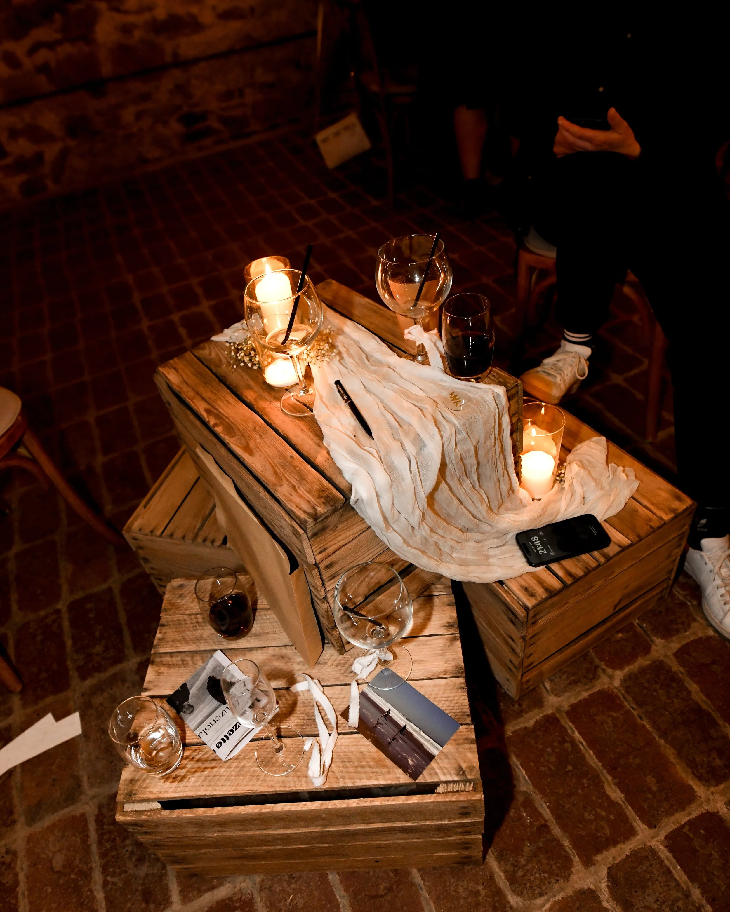 Une décoration de table chaleureuse avec des bougies allumées, des verres de vin rouge et blanc, des papiers et un stylo, sur une table en bois dans une ambiance intime et sombre.