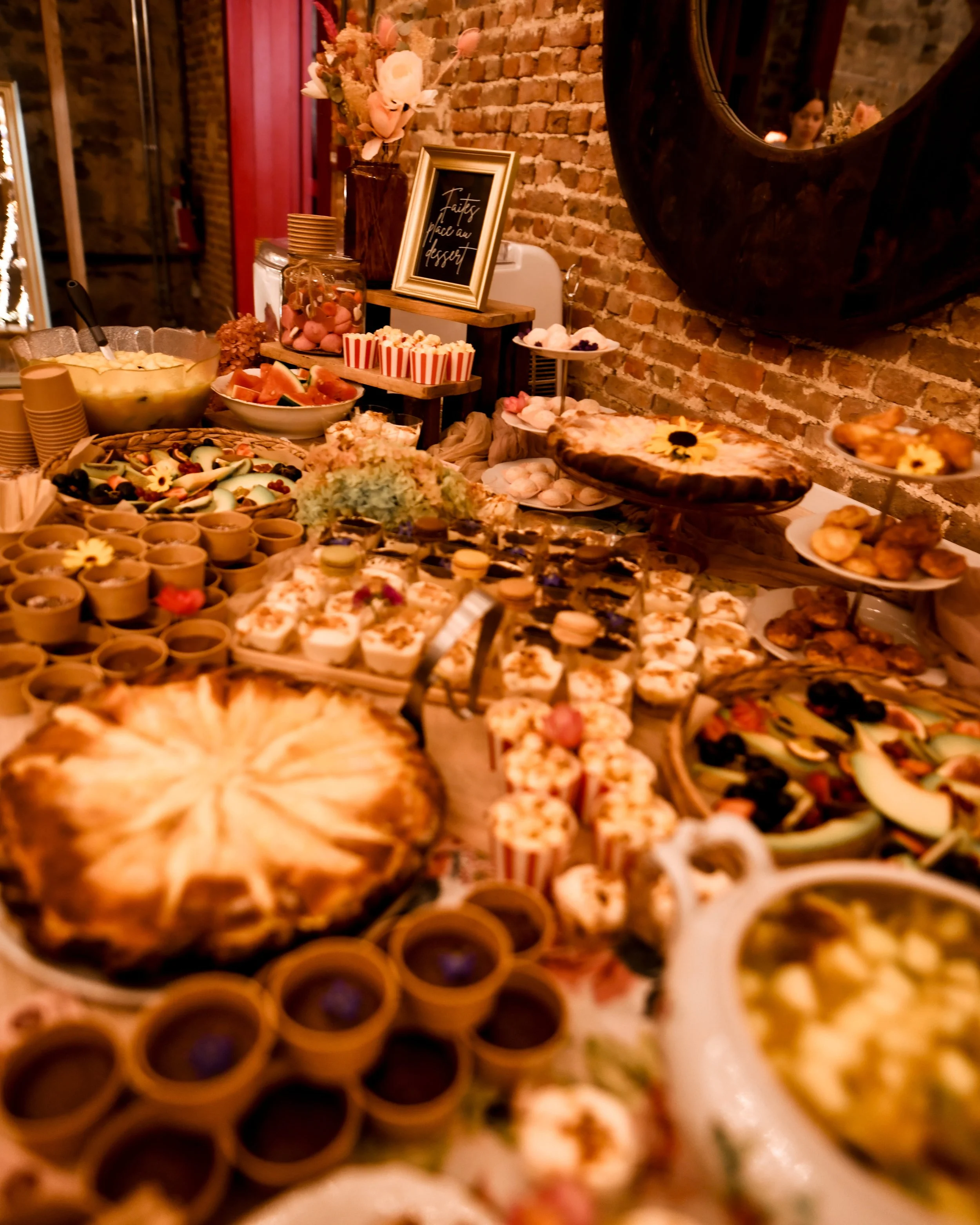 Buffet de desserts avec biscuits, tartes, cupcakes, fruits et fleurs, dans un décor chaleureux avec un mur en briques.