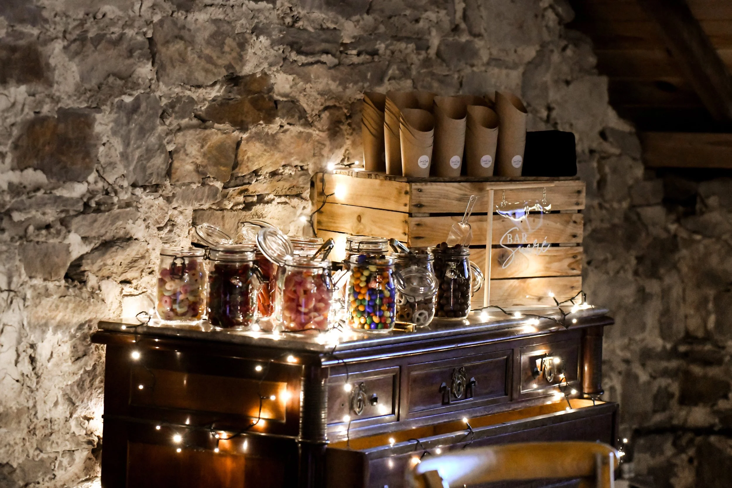 Une table en bois sombre décorée avec des bocaux remplis de bonbons colorés, sous un éclairage tamisé avec des guirlandes lumineuses, contre un mur en pierre.