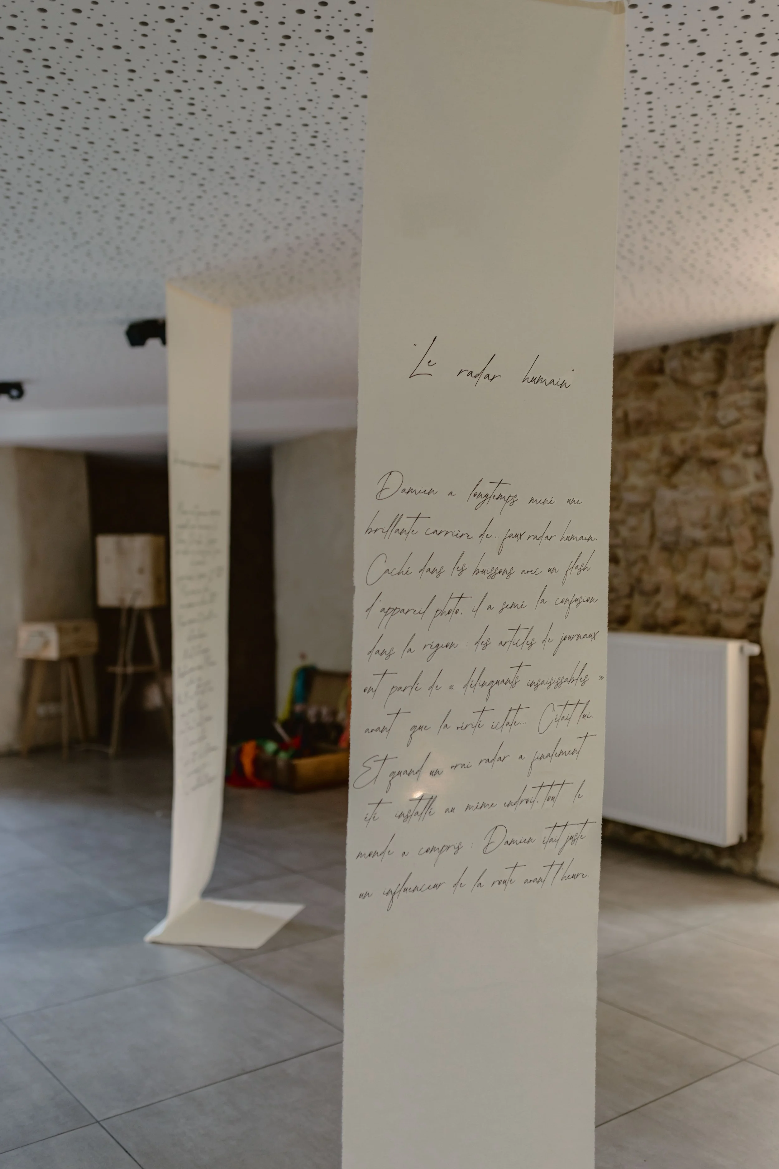 Une longue bande de papier écrit avec une écriture cursive, suspendue dans une pièce avec un mur en pierre et un plafond à motifs perforés.
