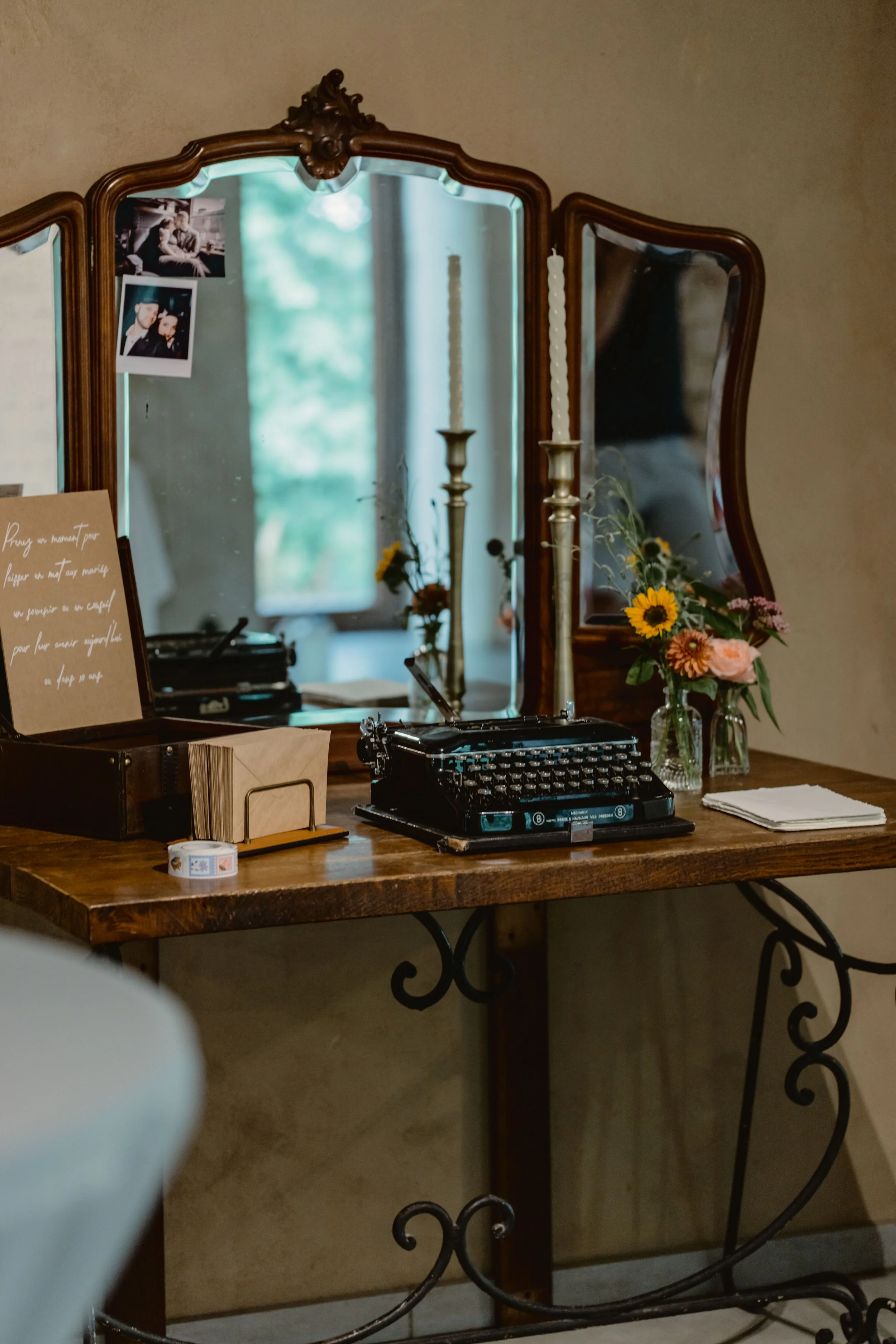 Un meuble vintage avec un grand miroir en bois, décoré de photos anciennes, un vase avec des fleurs, un carnet, une boîte à musique, une machine à écrire et une bougie.