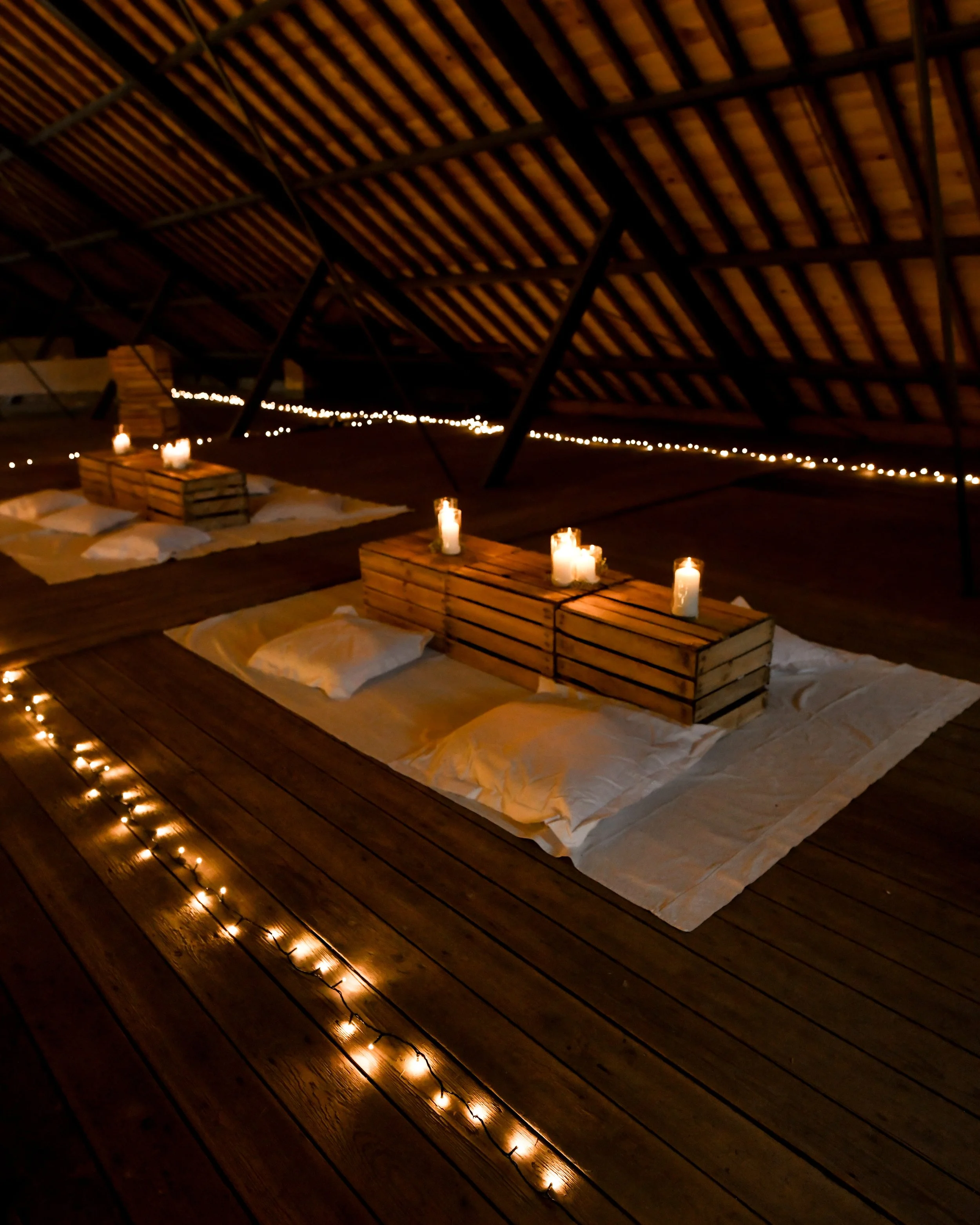 Une décoration intérieure chaleureuse avec des bougies allumées, des coussins blancs et des palettes en bois sur un sol en bois, sous un plafond en bois avec des poutres, ambiance ambiance cosy et romantique.