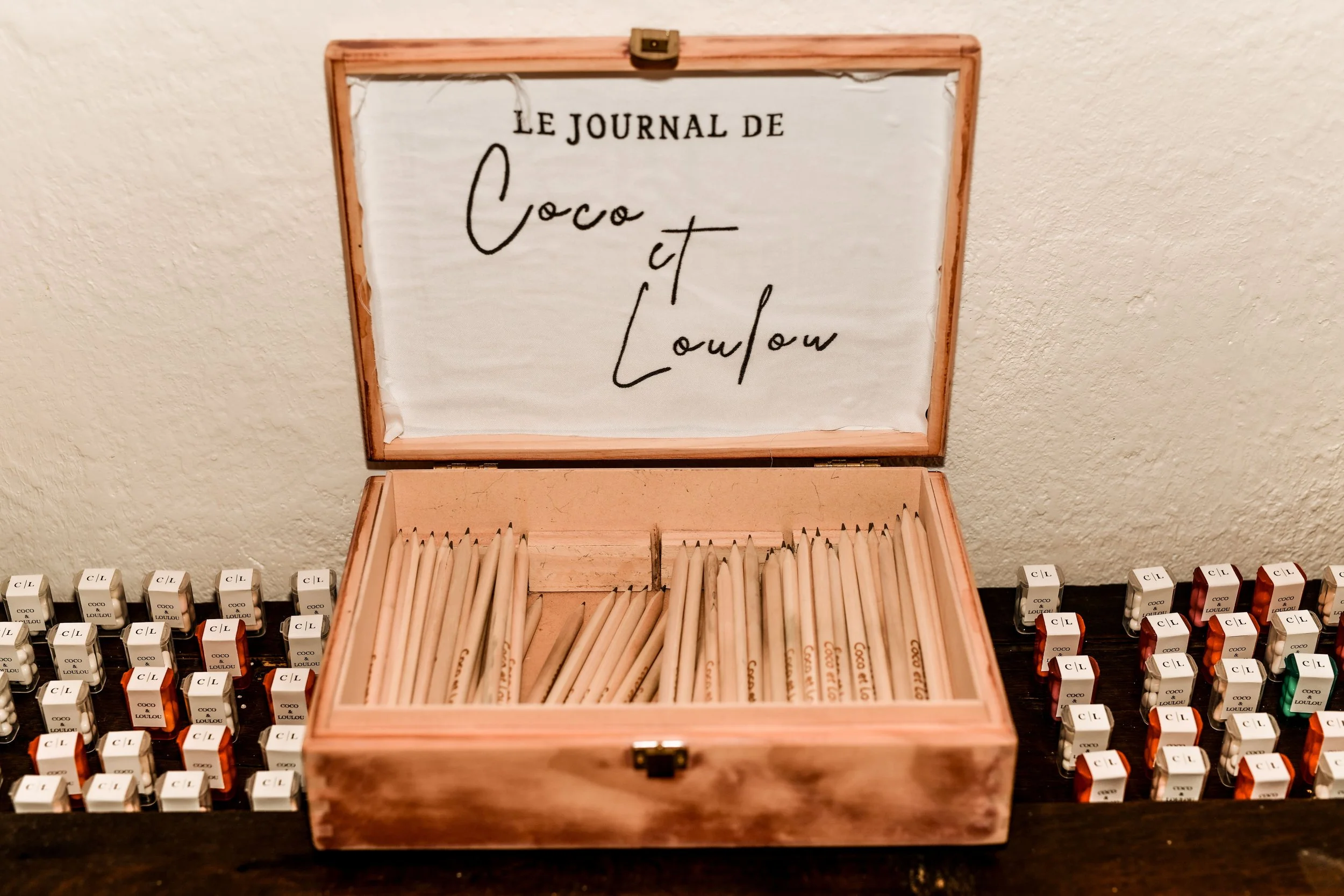 Une boîte en bois ouverte contenant des crayons ou des crayons de couleur, avec le fond d'une table en bois. En arrière-plan, il y a plusieurs petits flacons de peinture ou d'encre de différentes couleurs, tous disposés alignés.