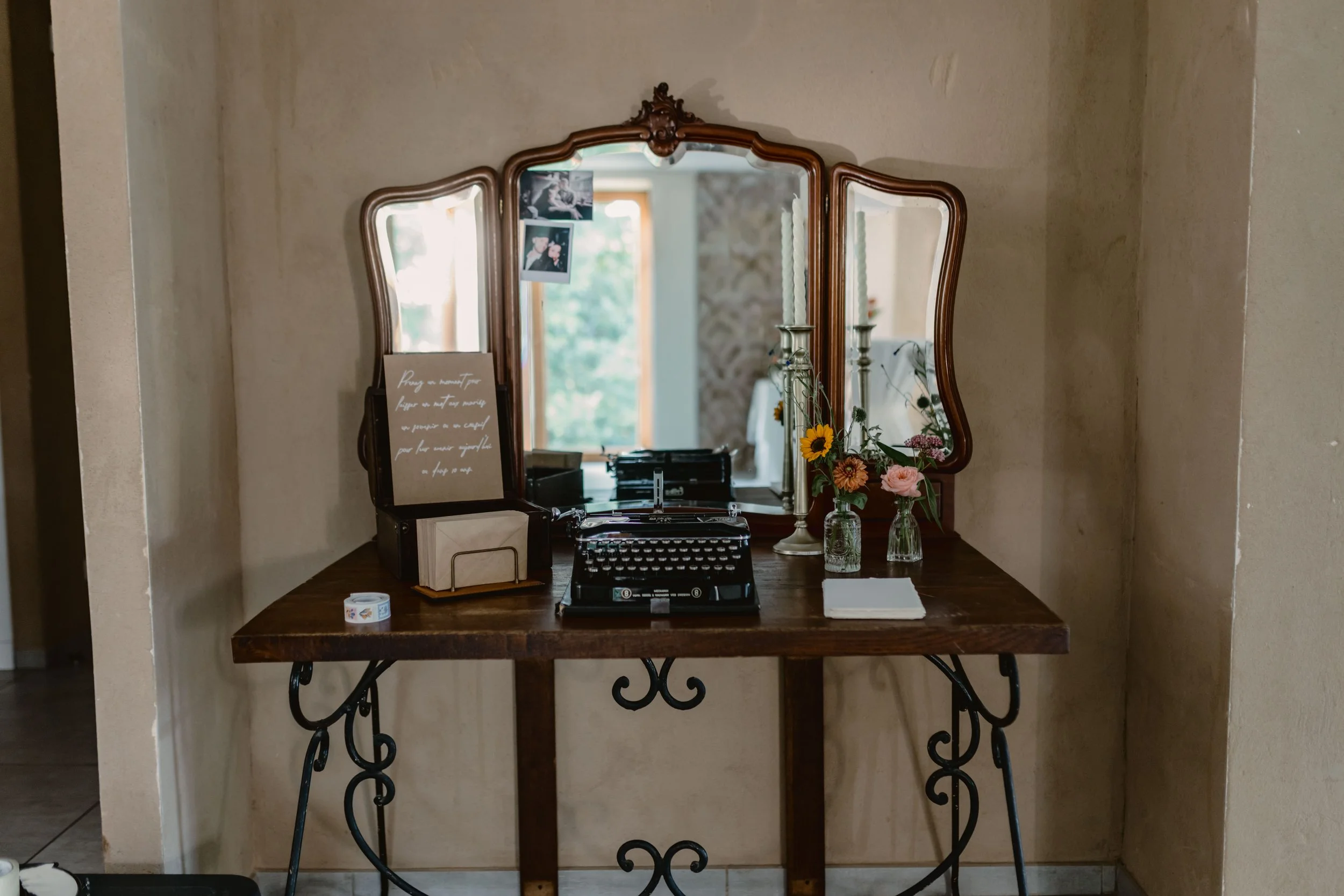 Miroir ancien avec cadre en bois, posé sur une console en bois, décoré de fleurs et de bougies, dans un intérieur lumineux.