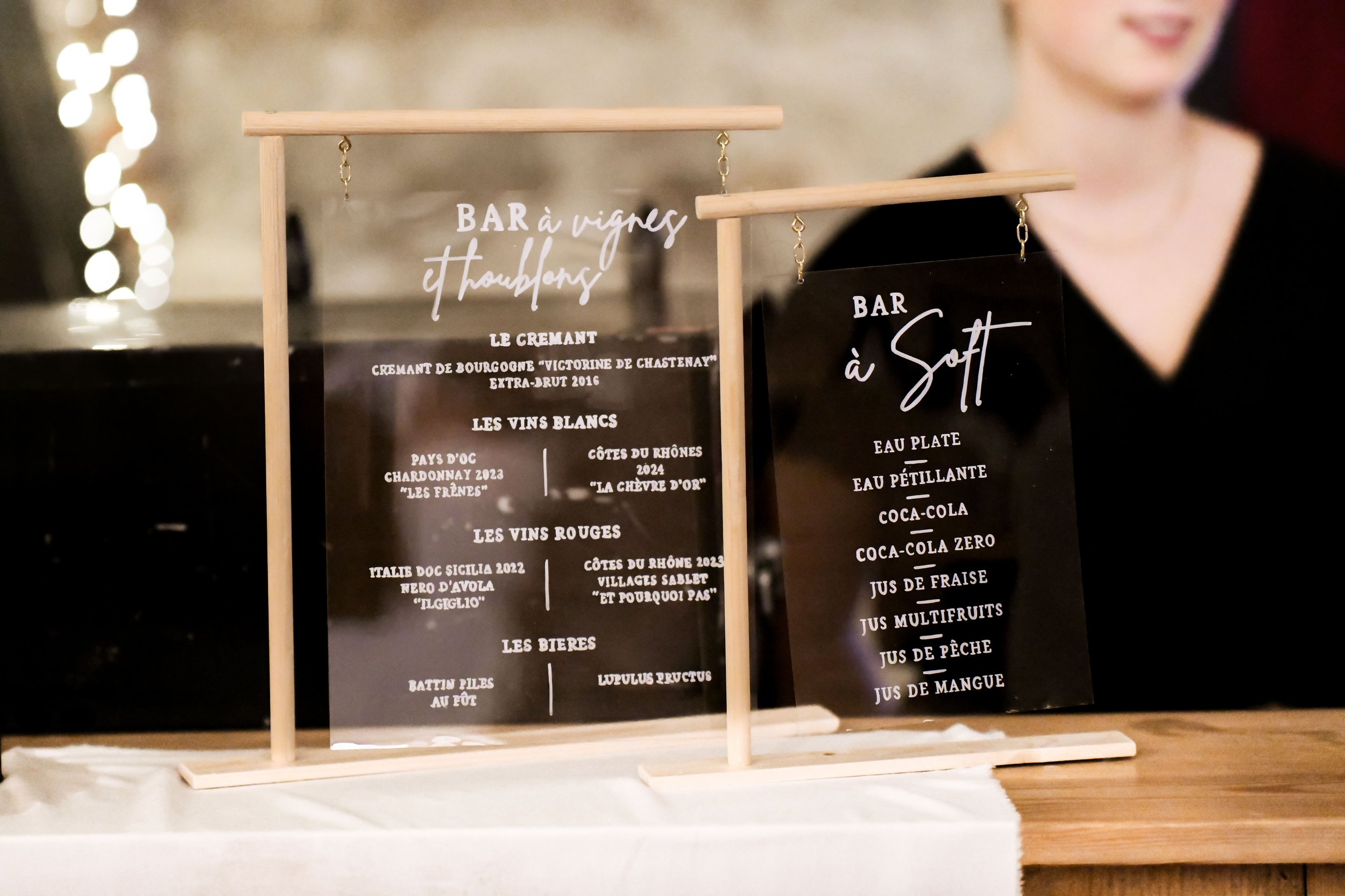 Deux panneaux noirs et en verre avec des inscriptions blanches, décorés de baguettes en bois, affichant une sélection de vins et de boissons dans un bar.