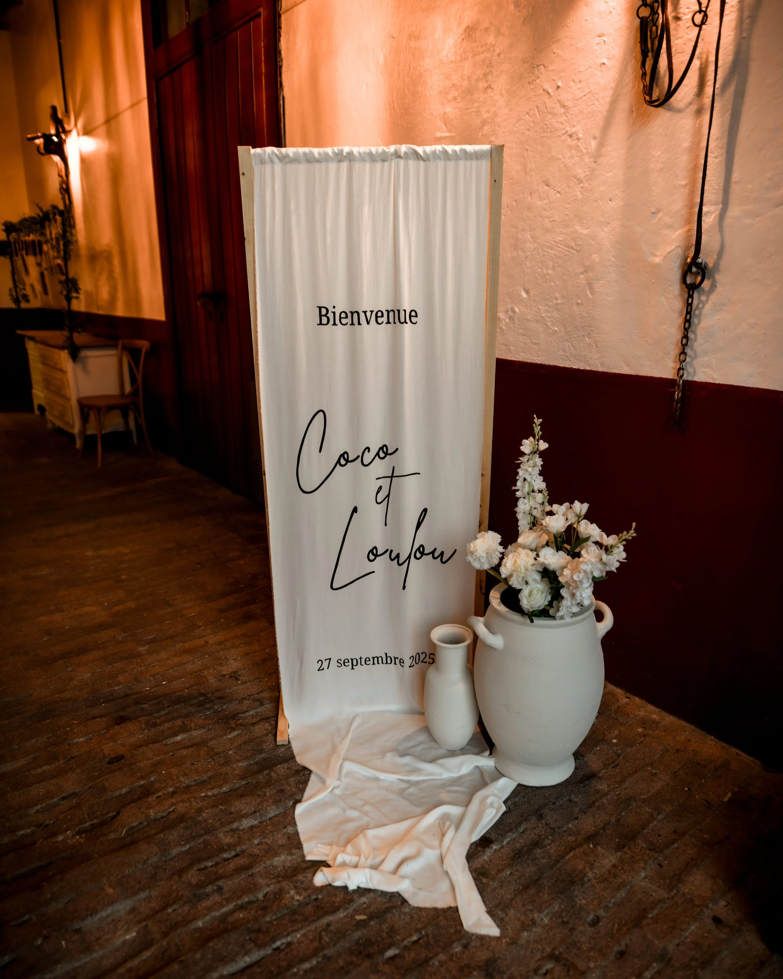 Panneau blanc avec texte en français pour un mariage, disant « Bienvenue Coco et Loulou » avec la date 27 septembre 2025, placé à côté de vases blanc remplis de fleurs blanches, dans une pièce avec murs en pierre et sol en bois.