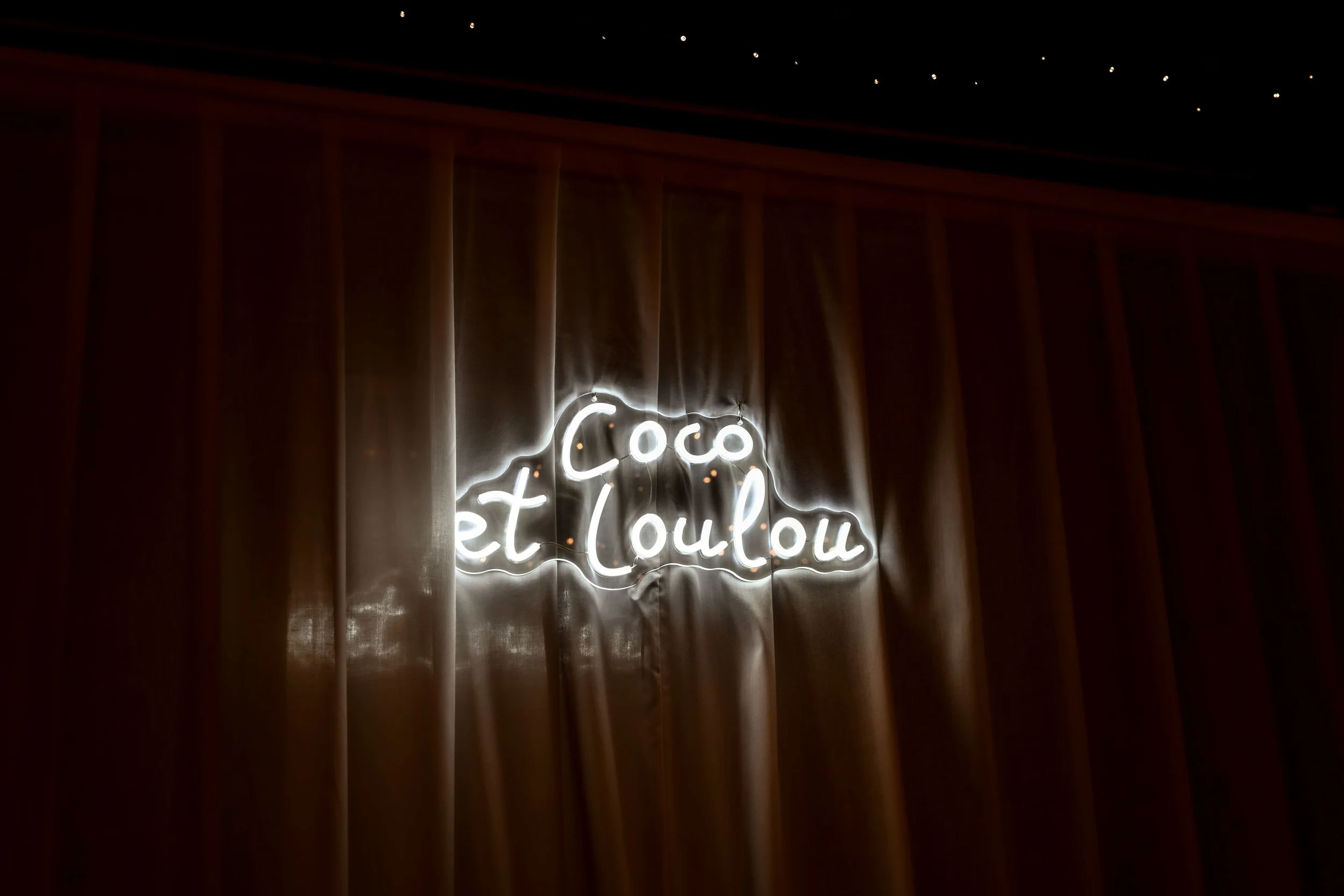 Un panneau lumineux en néon blanc avec la phrase en français "Coco et Loulou" sur un mur en bois, sous un plafond avec des petites lumières