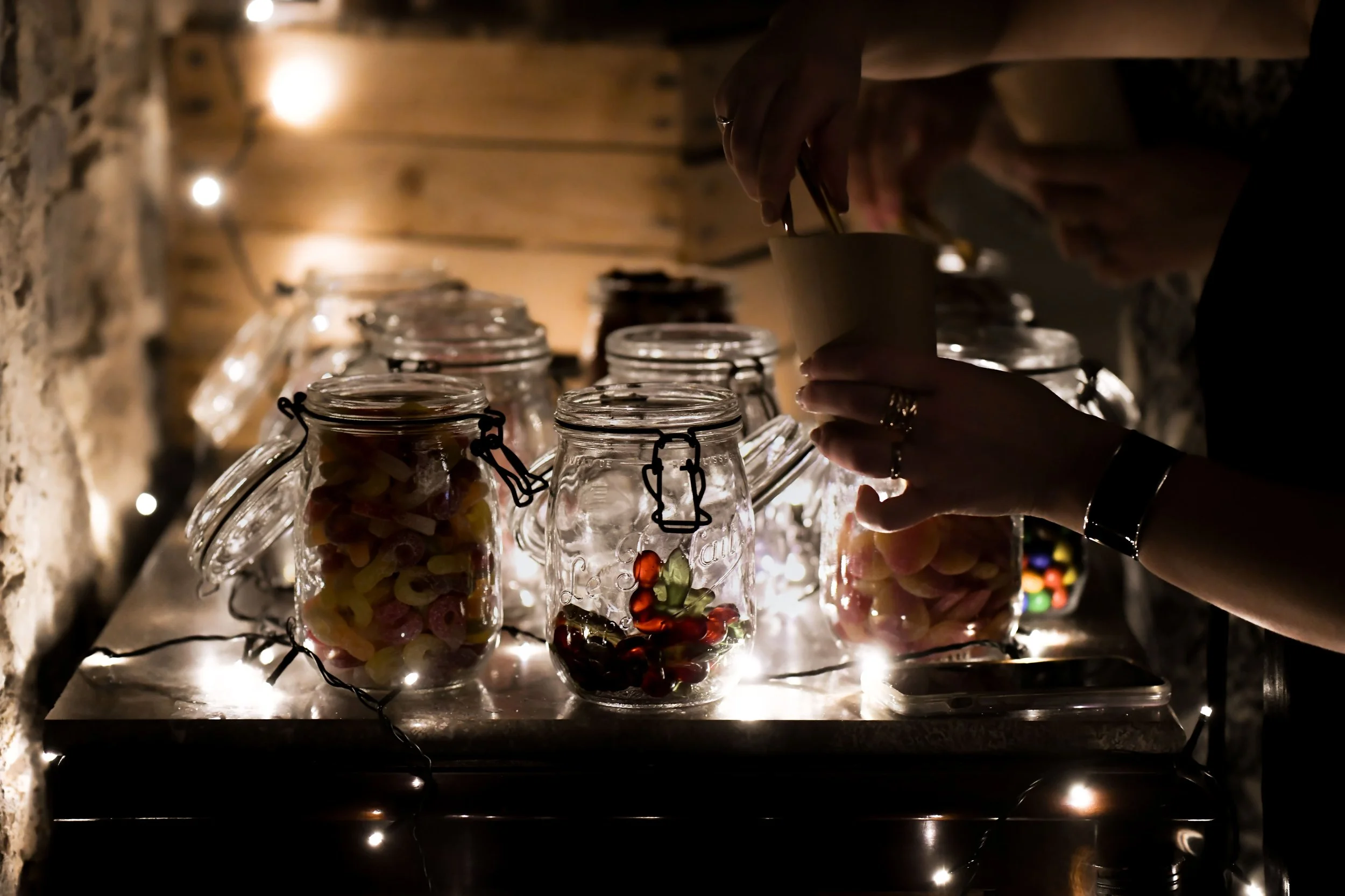 Une personne prenant des bonbons dans des bocaux en verre, éclairés par des guirlandes lumineuses, dans un décor chaleureux et rustique.