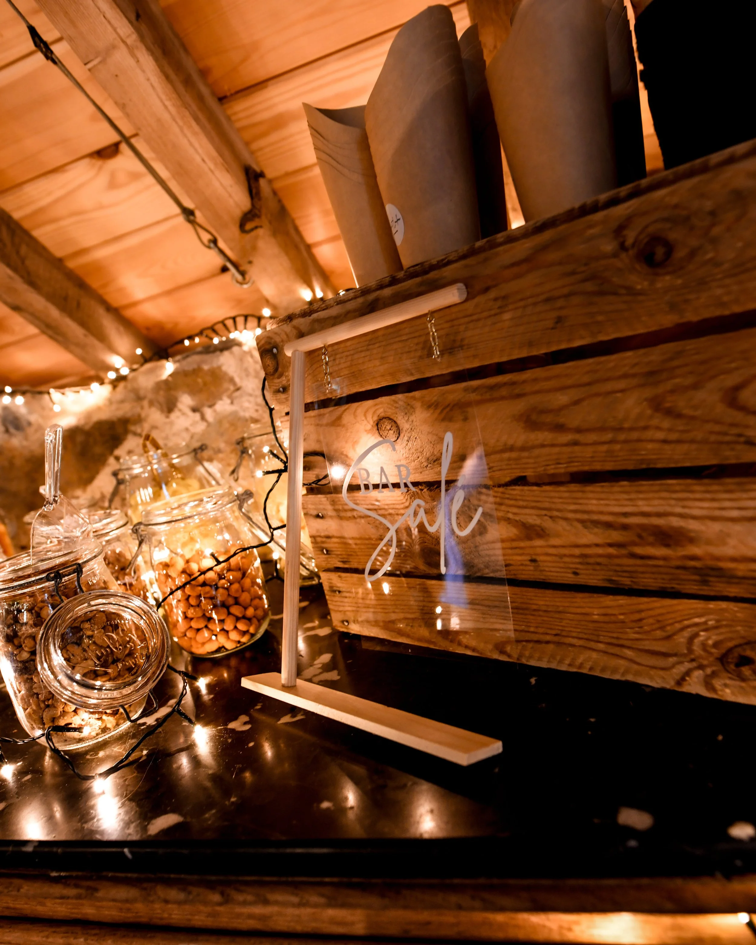 Décoration intérieure d'un bar avec un éclairage tamisé, comprenant un panneau en bois avec le mot 'Bar' écrit dessus, des bocaux en verre avec des aliments, et des guirlandes lumineuses