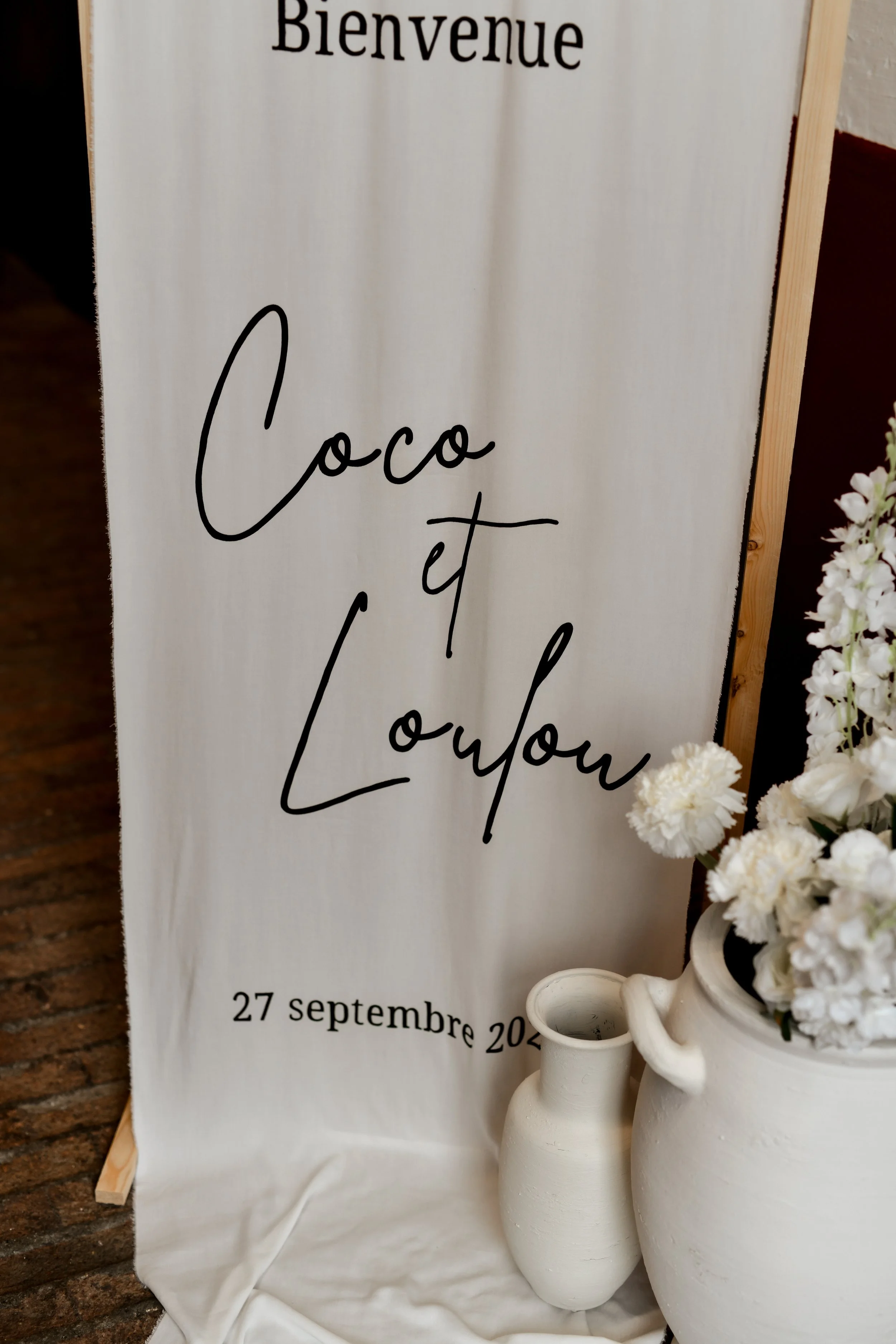 Une bannière avec le texte "Bienvenue Coco et Loulou" et la date "27 septembre 2025". À côté, des vases blancs contenant des fleurs blanches.