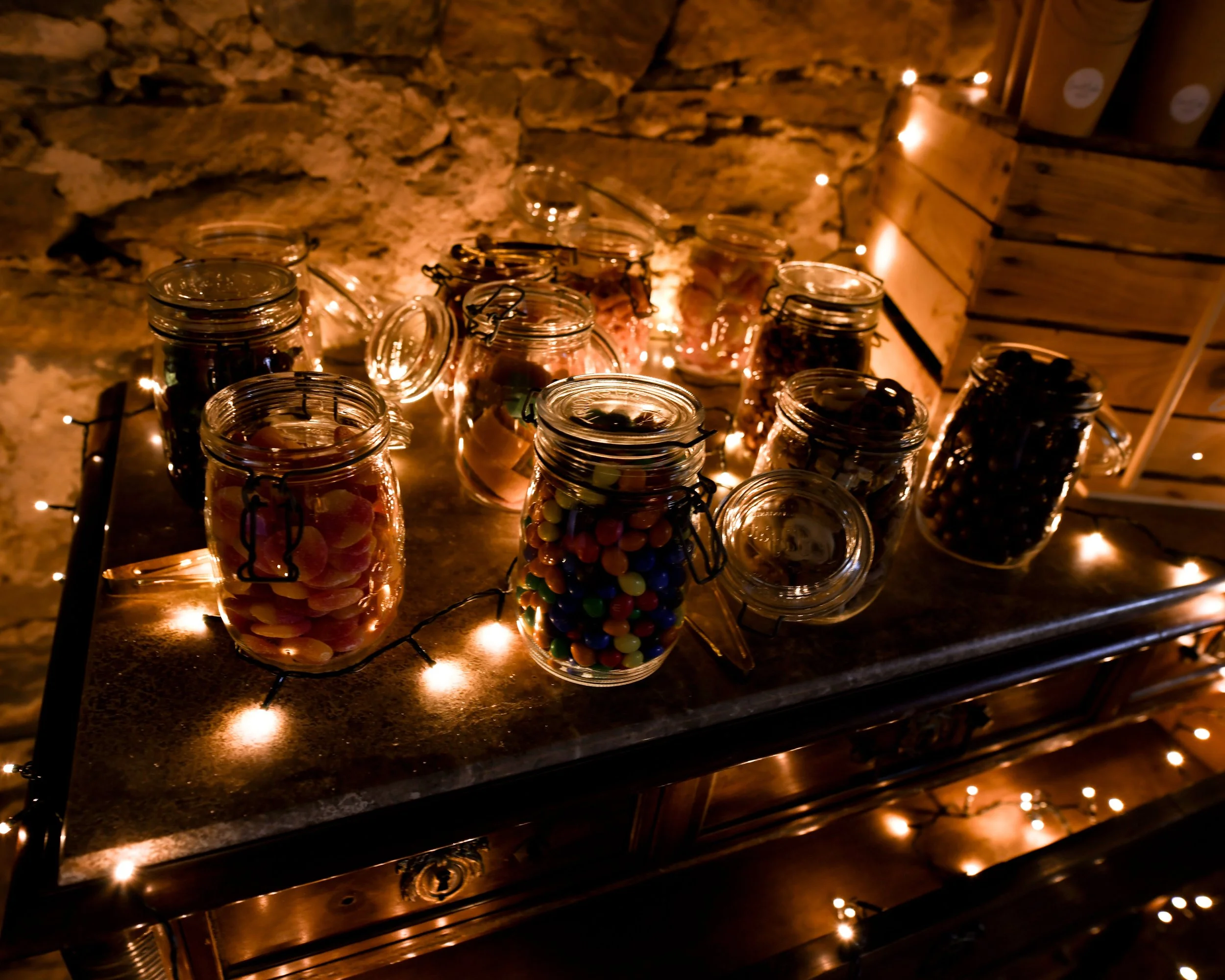 Verrines en verre remplies de bonbons colorés, éclairées par des guirlandes lumineuses, dans une ambiance chaleureuse et rustique.
