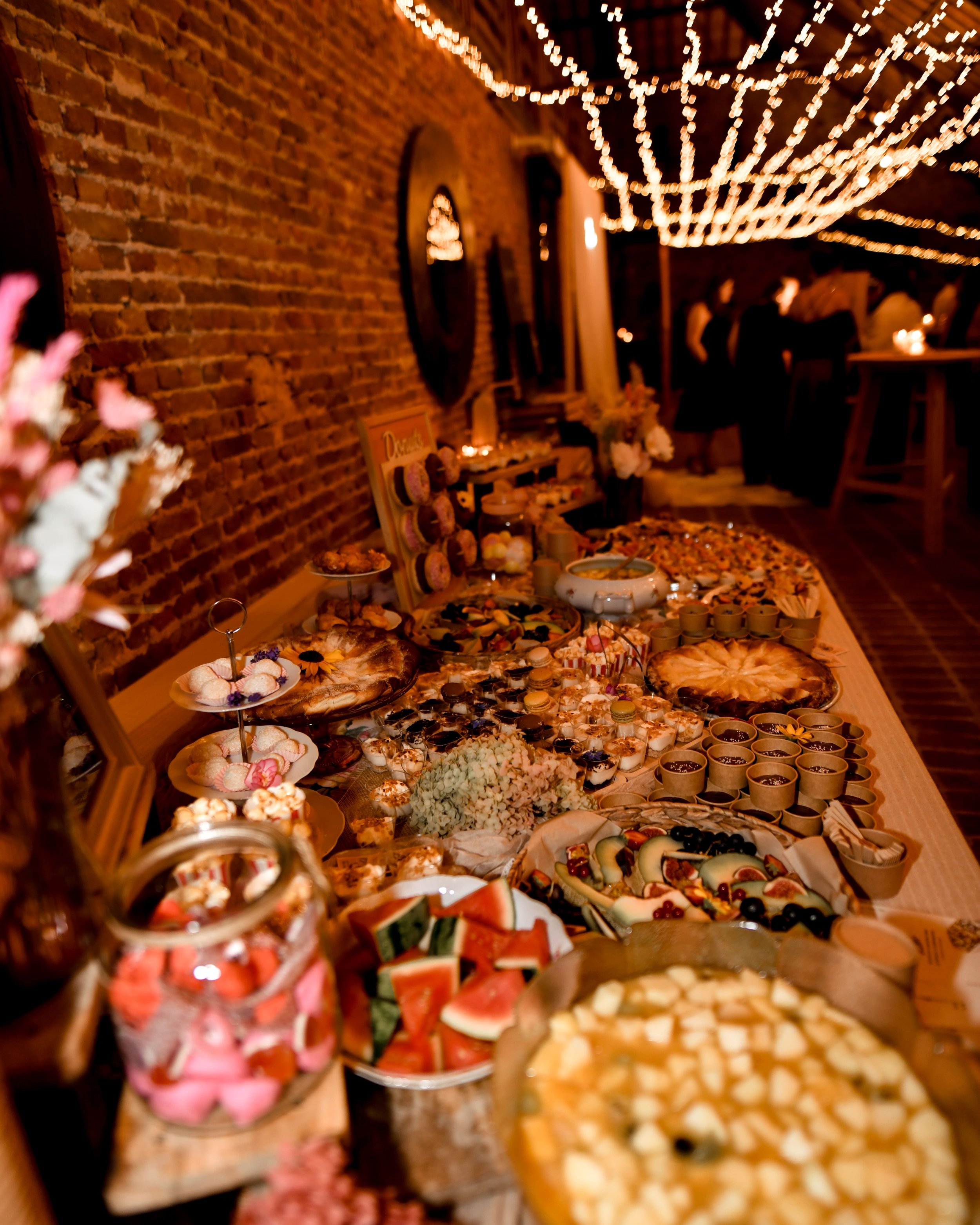 Un buffet de desserts et de fruits dans une ambiance chaleureuse avec des guirlandes lumineuses. Des petits fours, macarons, fruits coupés et autres douceurs disposés sur une table en bois, dans une pièce à murs en briques apparentes.