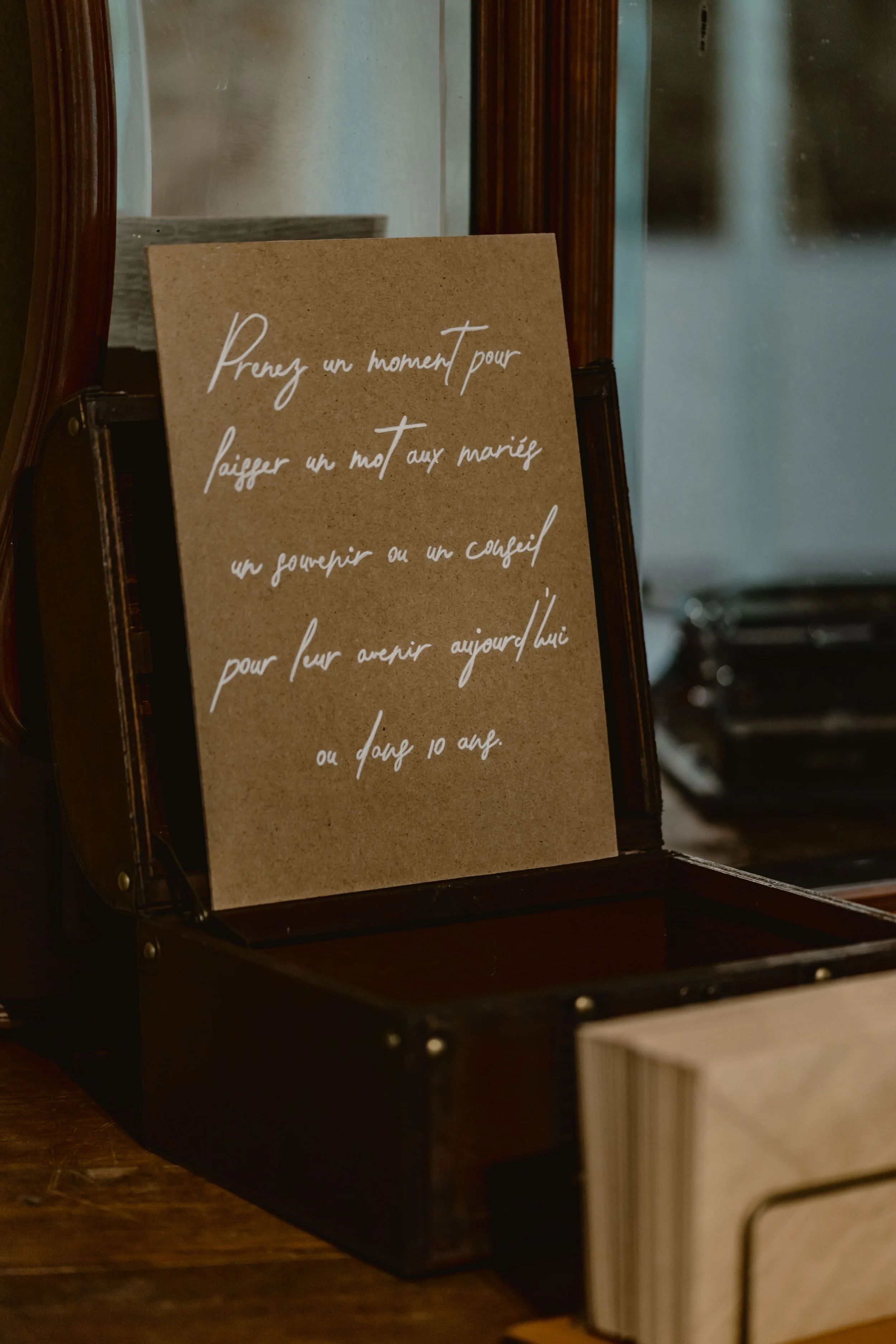 Une affiche en papier brun avec une écriture manuscrite blanche posée dans un cadre en bois, placé dans une boîte en bois foncé. La réflexion sur la vitre indique une présence derrière le photographe.