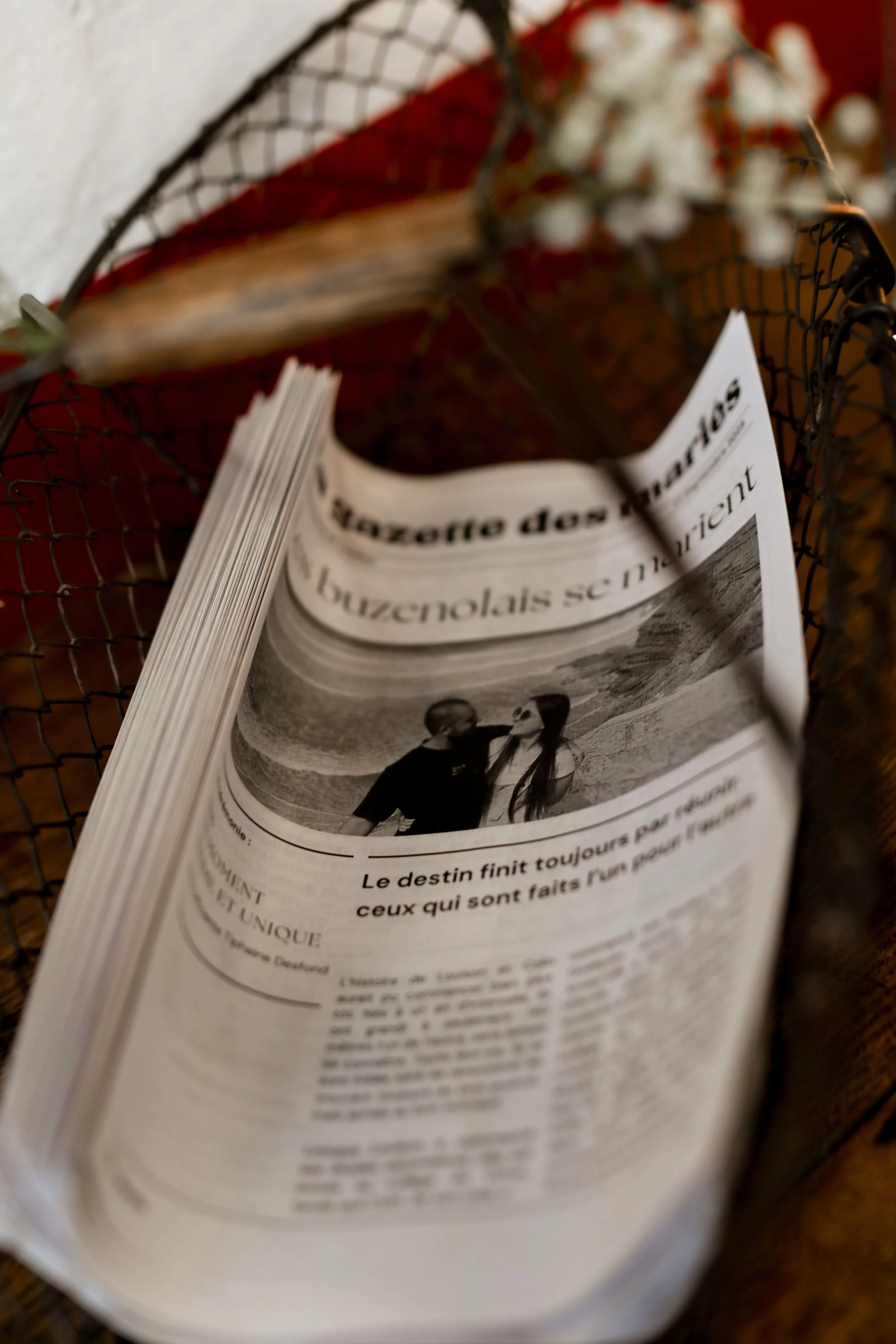 Un journal plié avec une photo en noir et blanc d’un couple à la plage, entouré d’un panier en métal avec des fleurs blanches