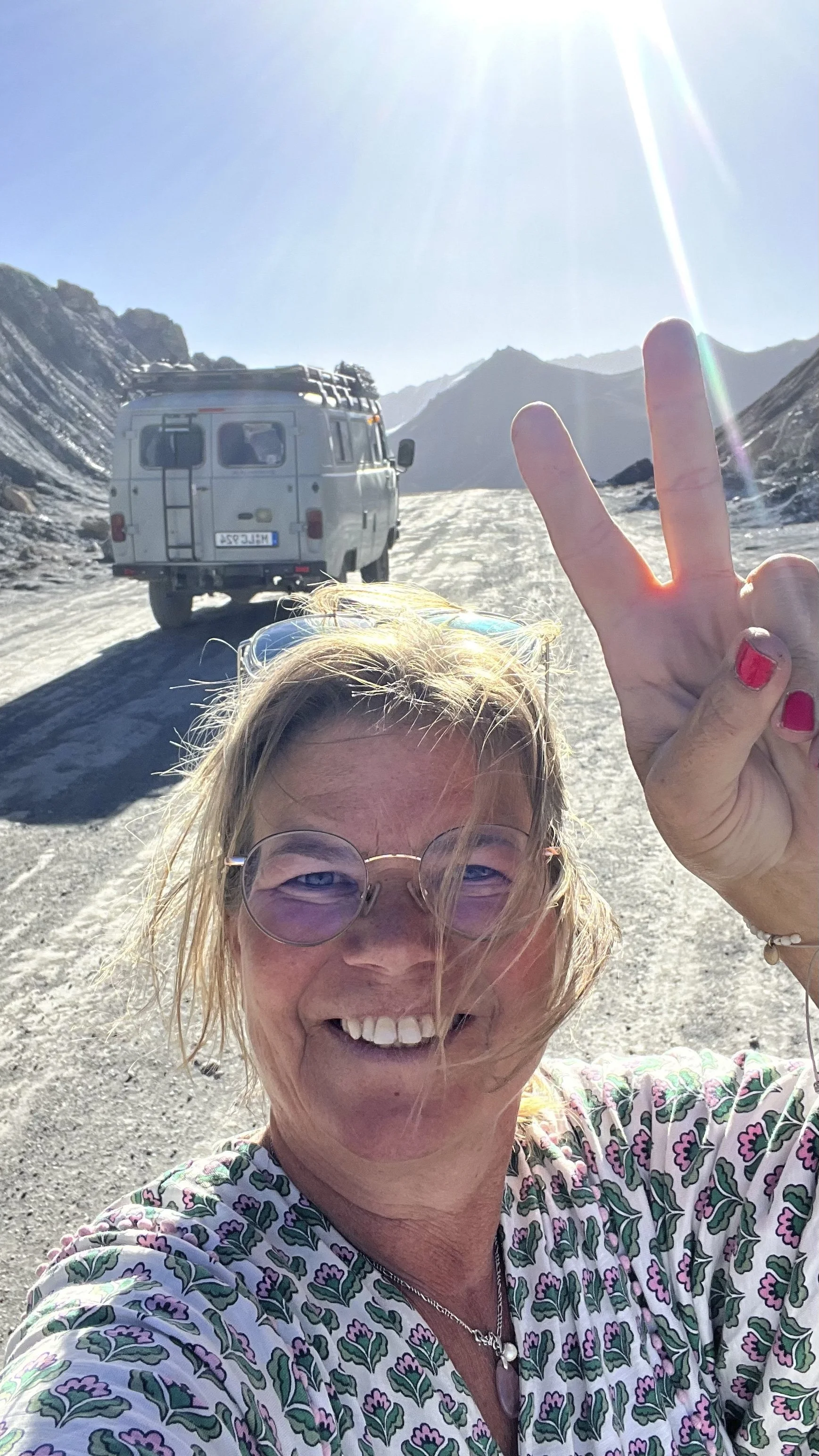 Eine lachende Frau mit Sonnenbrille macht ein Selfie vor einem weißen Van in einer bergigen, schneebedeckten Landschaft bei Sonnenlicht, während sie peace-Zeichen zeigt.