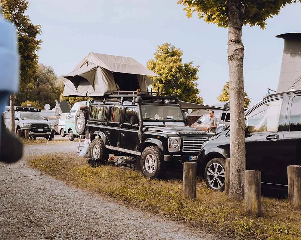 Ein schwarzer Land Rover Defender mit einem Dachzelt auf dem Dach, parkend auf einem Campingplatz bei Bäumen, umgeben von anderen Fahrzeugen.