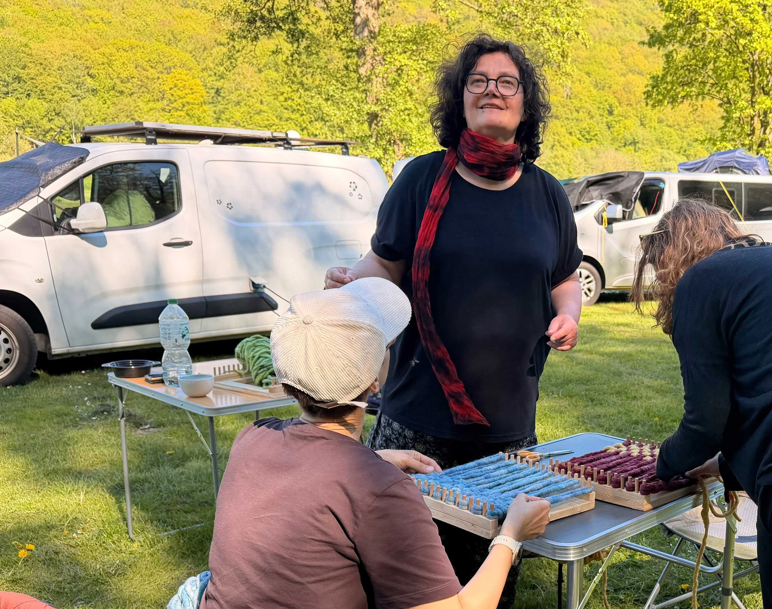 Menschen, die im Freien an einem Tisch mit einem Modell eines Webstuhls arbeiten, während andere Personen im Hintergrund sitzen.