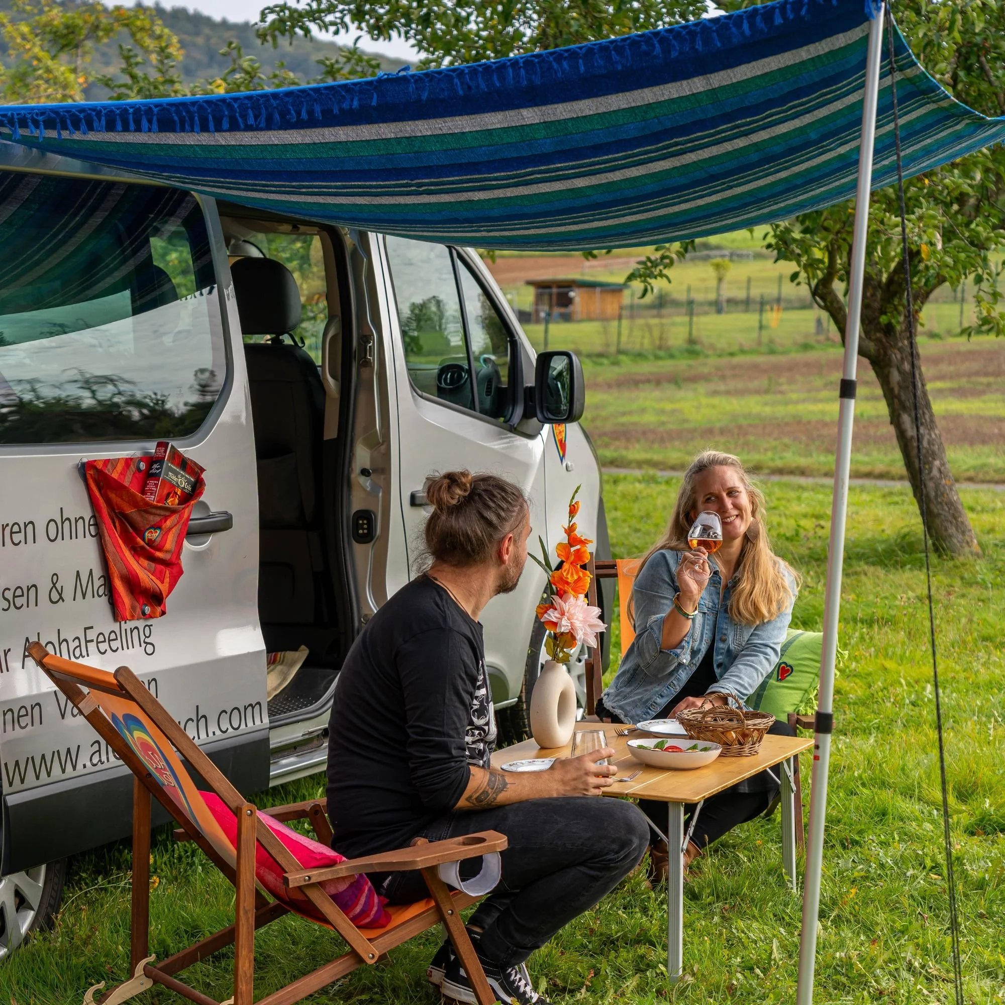 Menschen sitzen bei einem Picknick im Freien vor einem umgebauten Van, der einen Sonnenschutz aus buntem Stoff hat. Es gibt Getränke und Blumen auf dem Tisch, und die Umgebung ist grün mit Bäumen und Wiesen.