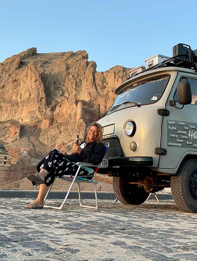 Frau sitzt in einem Klappstuhl neben einem Campingbus in einer bergigen Landschaft und hält ein Getränk in der Hand, Sonnenuntergang im Hintergrund.