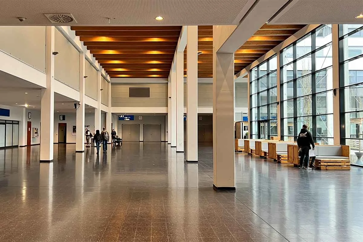 Innenansicht eines modernen Flughafenterminals mit großen Fenstern, Holzbalkendecke und einigen wartenden Passagieren.