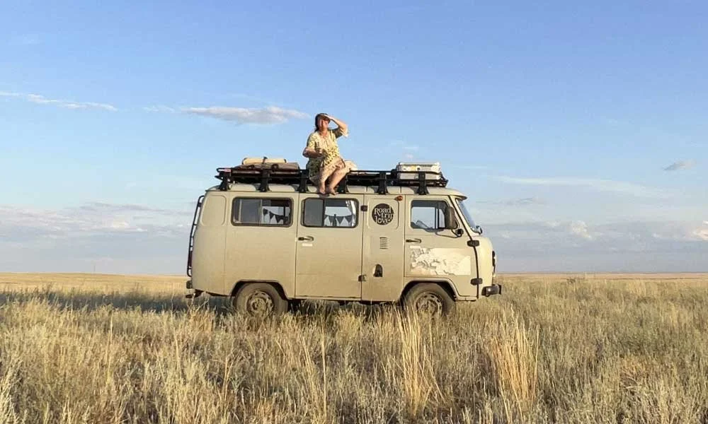 Frau sitzt auf dem Dach eines alten Vans in einer offenen Graslandschaft, blickt in die Ferne, bei Sonnenschein.