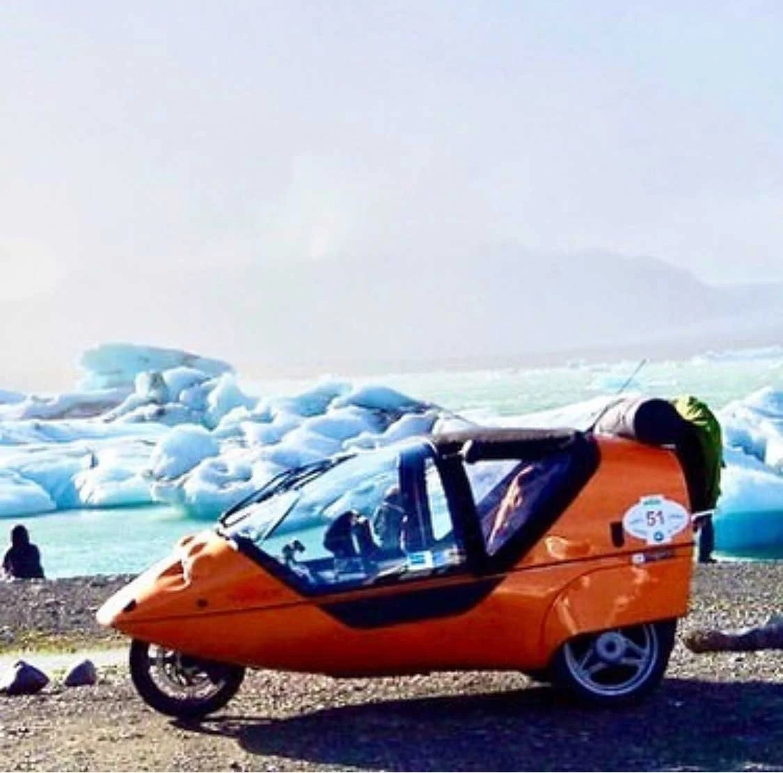 Ein orangefarbenes Fahrzeug mit einem geschlossenen Cockpit auf einer felsigen Küste vor einem Gletscher