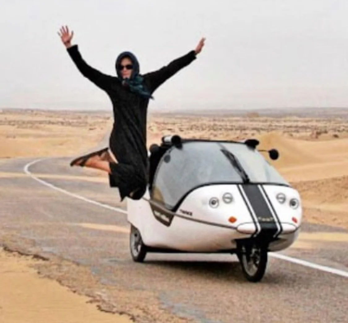 Person auf einem Einrad, das wie ein kleines Auto aussieht, in einer Wüstenlandschaft. Der Mensch trägt Sonnenbrille, schwarzen Rock, schwarzen Oberteil, Schal und ist lachend mit ausgestreckten Armen.
