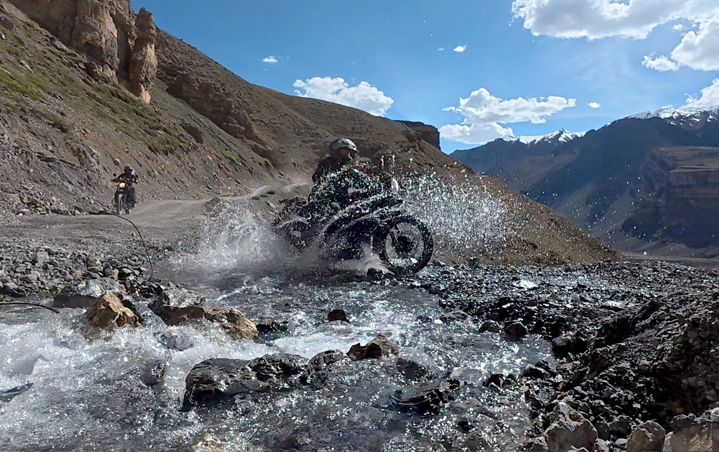 Motorradfahrer durchquert einen Fluss in einer Berglandschaft, während ein anderer Motorradfahrer im Hintergrund fährt. Es ist sonnig mit einigen Wolken am Himmel, die Berge sind schneebedeckt.