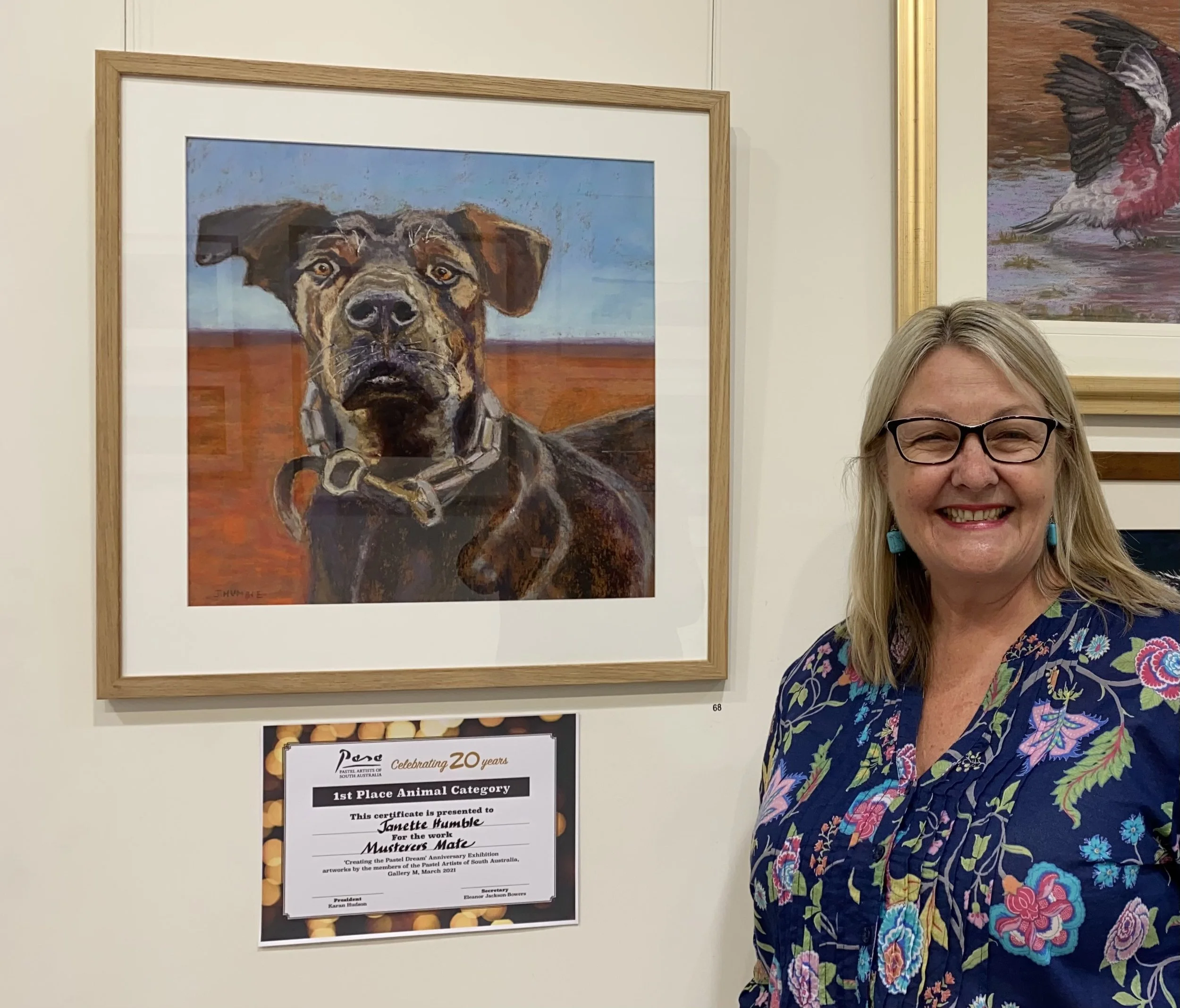 Janette Humble smiling and wearing glasses, standing next to a framed painting of a dog with a desert landscape background.