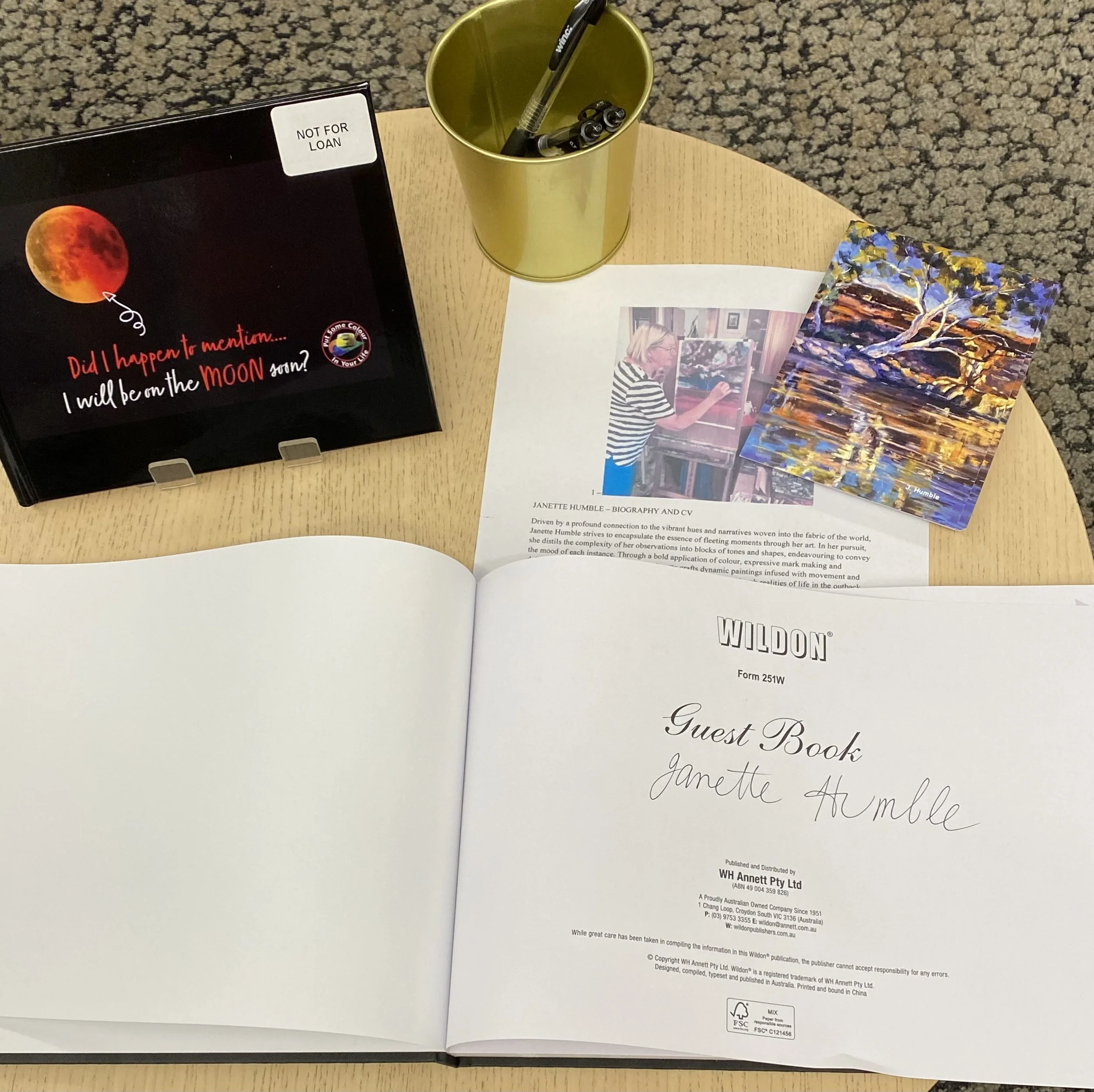 A wooden table with an open guest book labeled 'WILDON', some printed papers, a picture of an artist painting, an artwork of a sunset landscape, and a yellow cup holding pens. Additionally, there is a black sign with a lunar eclipse image and the text 'Did I happen to mention... I will be on the MOON soon?'