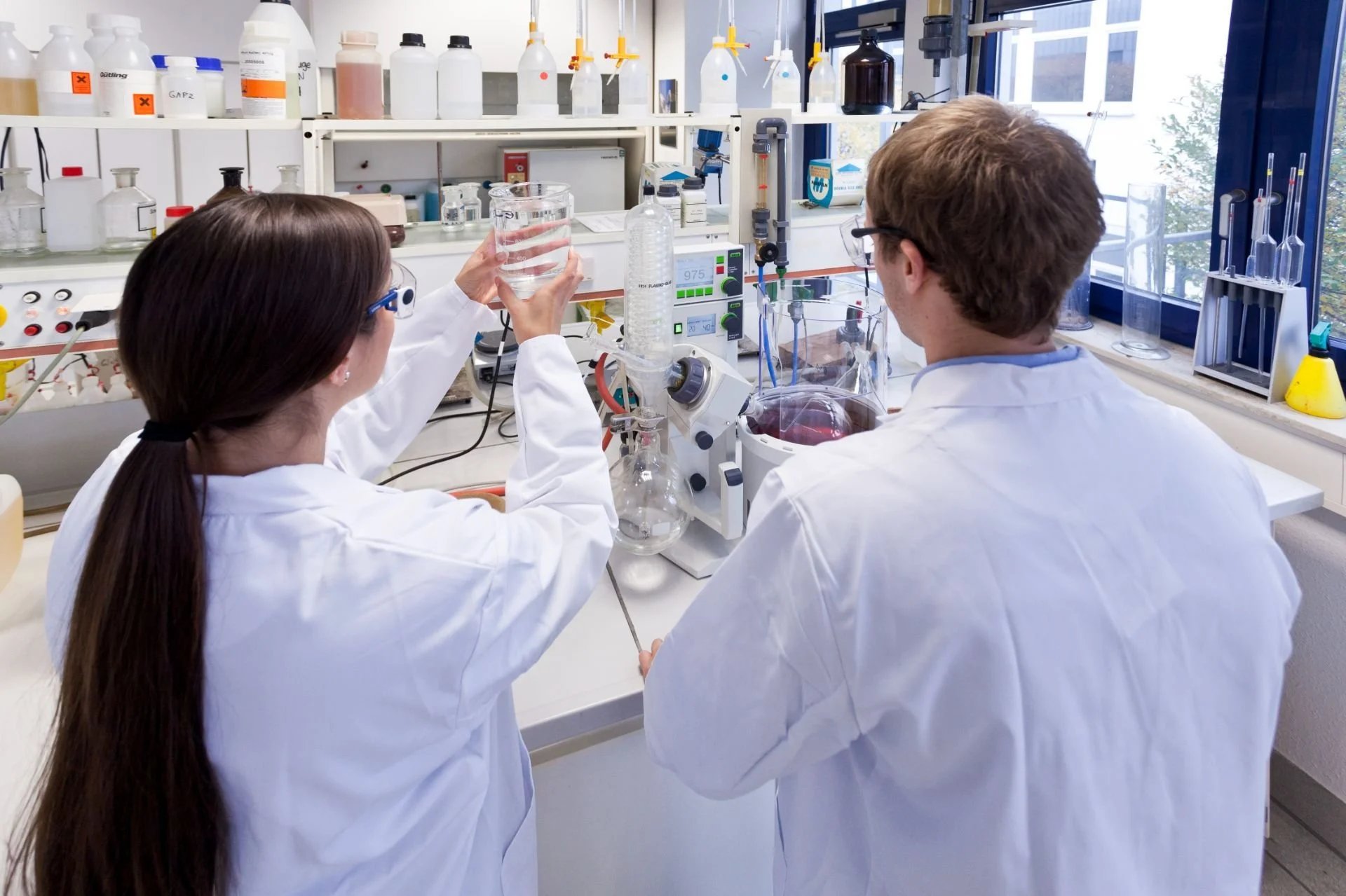 Zwei Wissenschaftler in Laborjackets arbeiten mit chemischen Substanzen in einem Labor mit mehreren Gläsern, Reagenzgläsern und Laborgeräten.