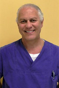 Ein älterer Mann in blauer medizinischer Kleidung vor einem hellgelben Hintergrund, lächelnd.