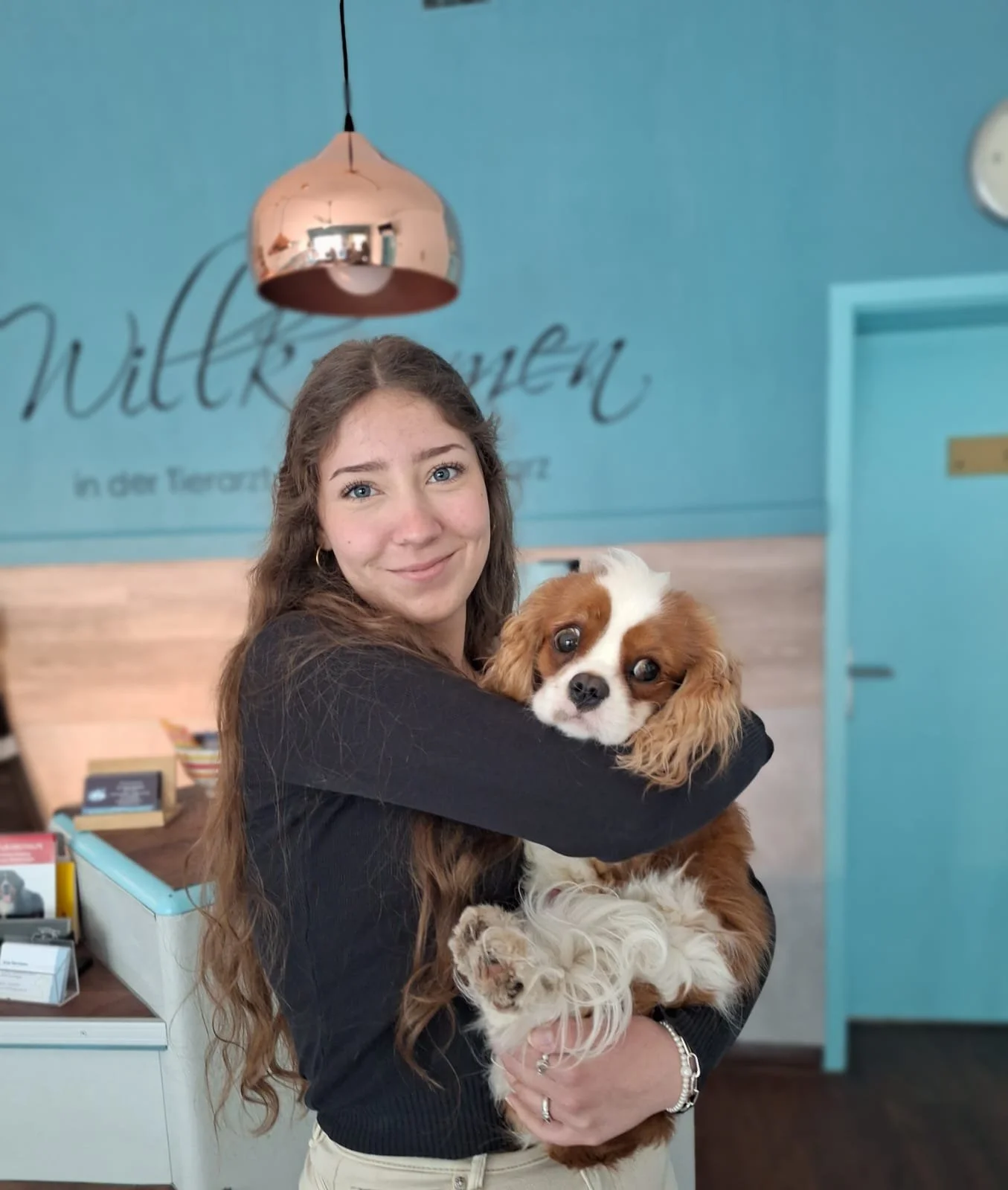 Eine junge Frau mit langen braunen Haaren hält einen Cavalier King Charles Spaniel in den Armen und lächelt in die Kamera. Sie befindet sich in einem Raum mit blauer Wand im Hintergrund, auf der Worte geschrieben sind. Es gibt eine Deckenlampe und eine Empfangstheke mit Broschüren.
