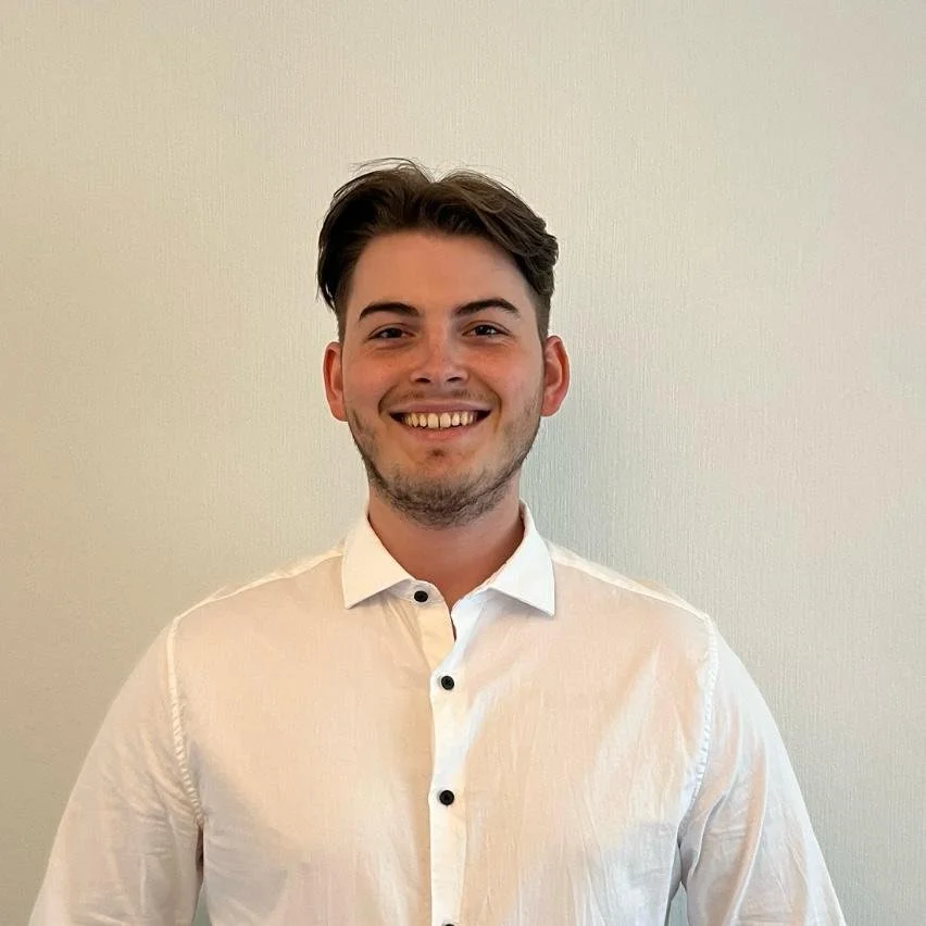 Ein junger Mann mit braunen Haaren und Bart, trägt ein weißes Hemd, lächelnd vor einem neutralen Hintergrund.
